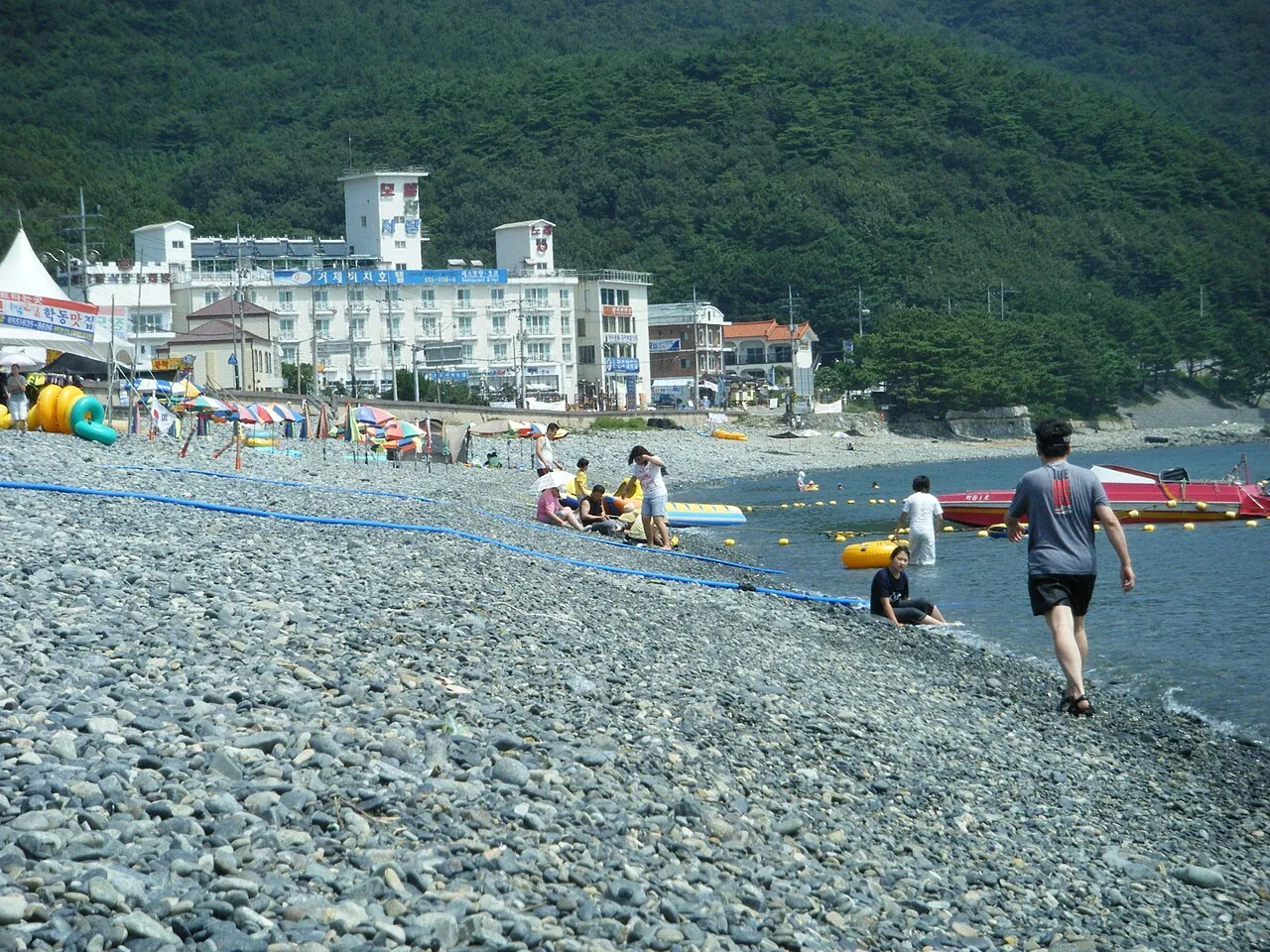 Swimmers and walkers on a grey pebble beach with hotels and wooded hills behind
