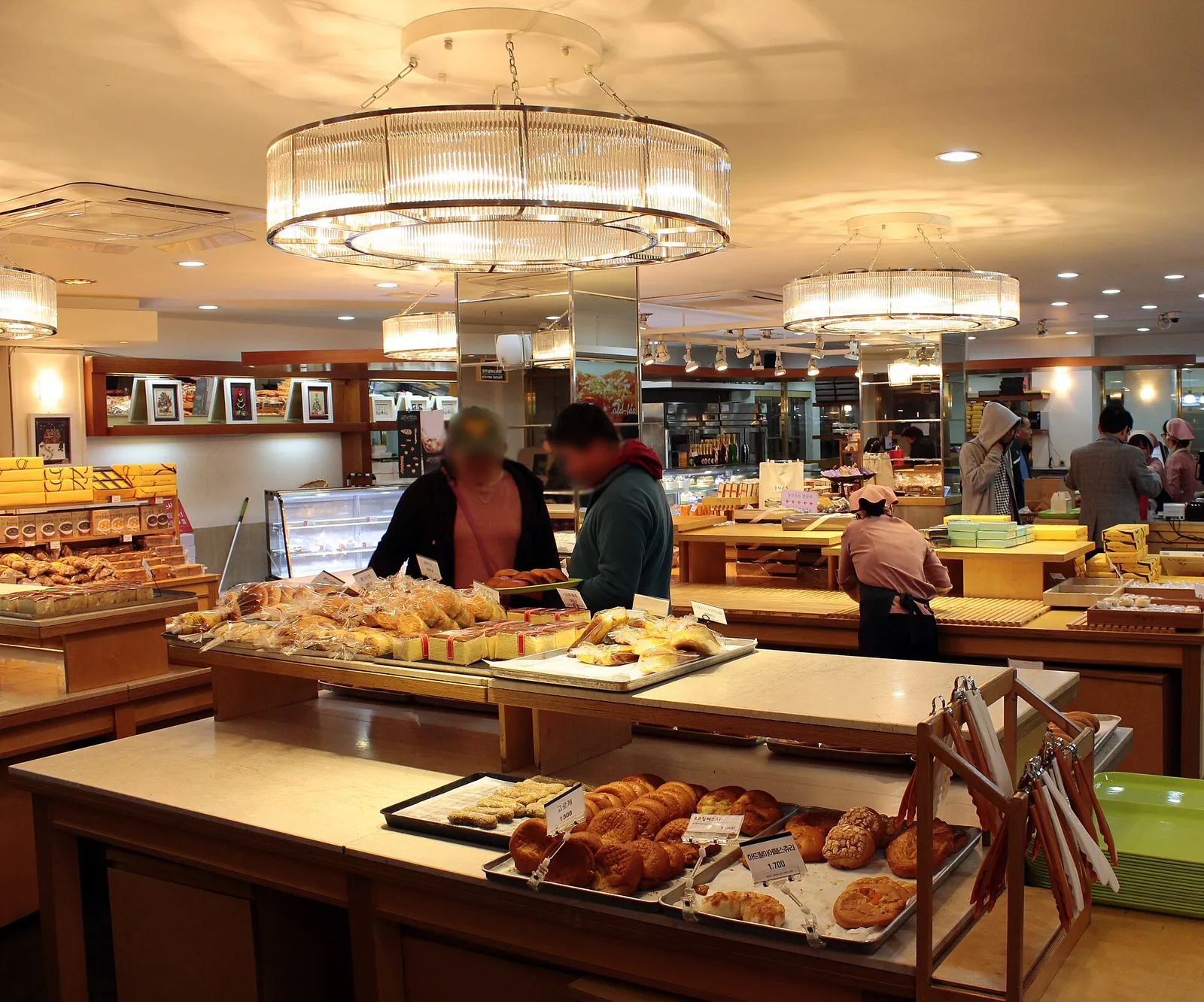 Shoppers choosing buns from trays on wooden tables under large round lamps in a bakery
