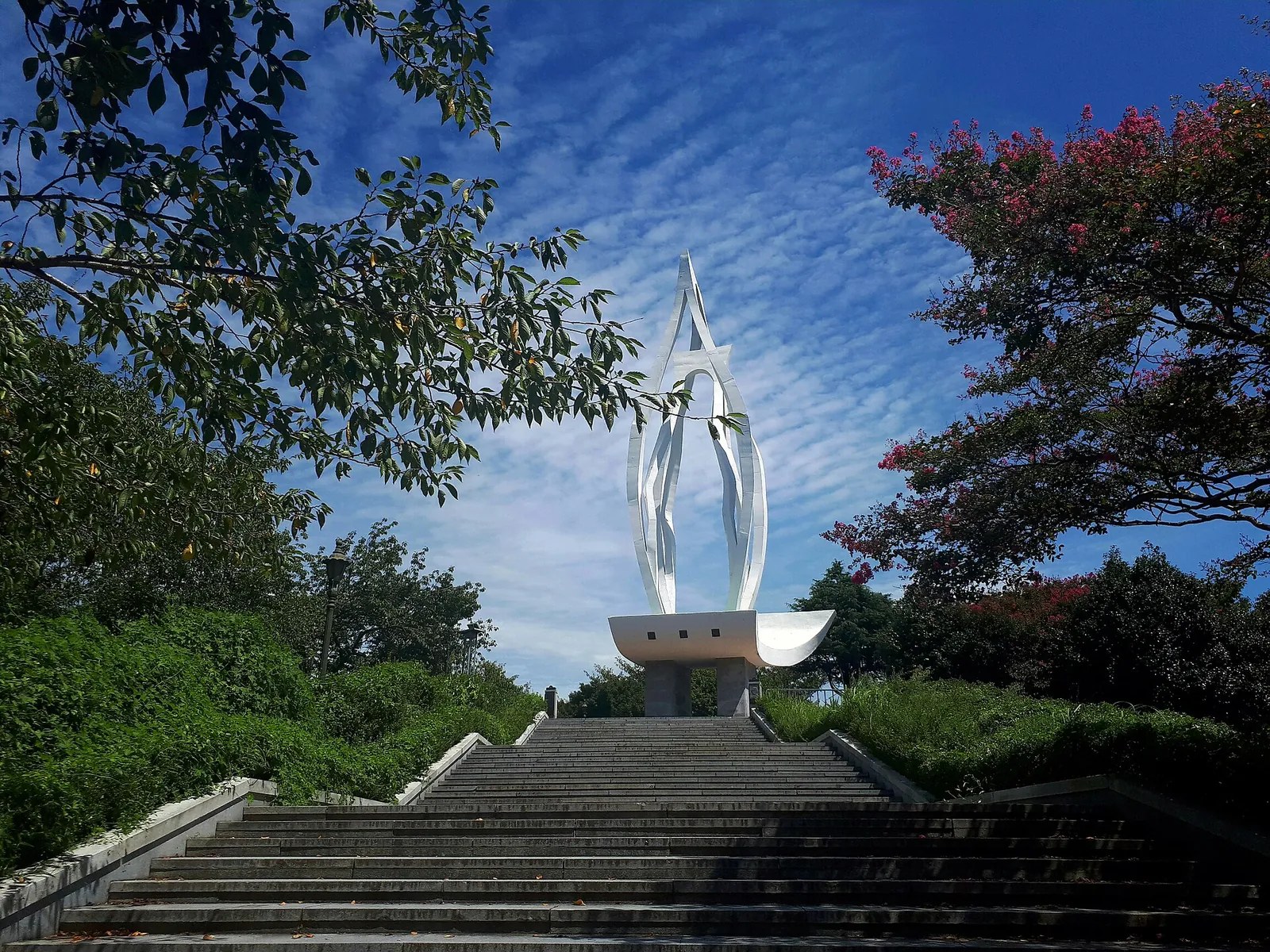 White flame-shaped tower at the top of stone steps, framed by trees against a blue sky