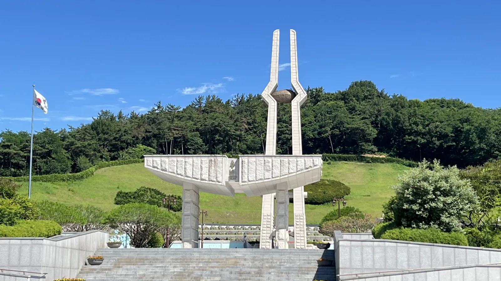 The white twin-pillared memorial tower at the May 18th National Cemetery in Gwangju under a blue sky, with wooded hills behind