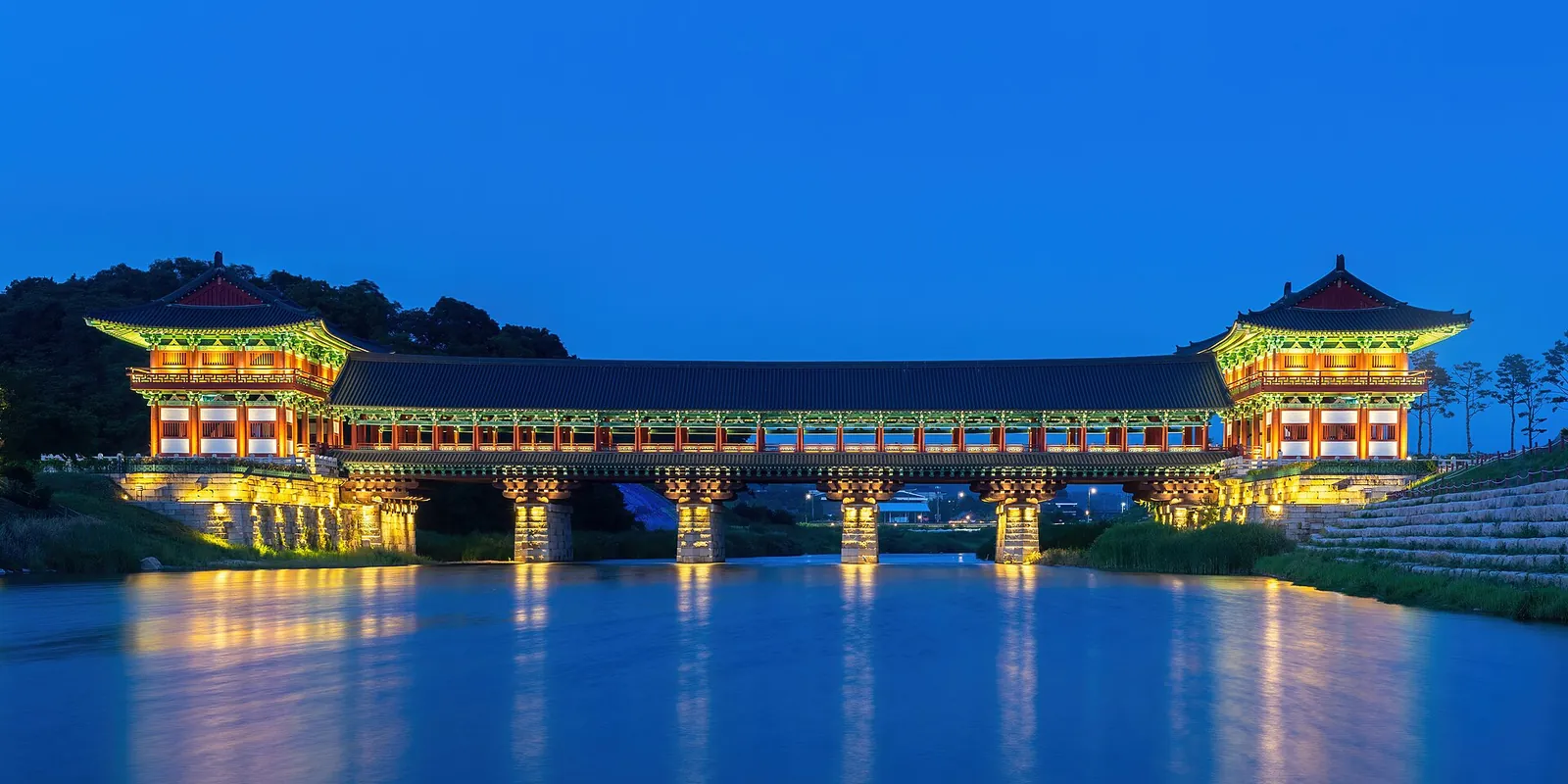 Woljeonggyo, a long roofed wooden bridge with gate towers, illuminated over the Namcheon stream at blue hour