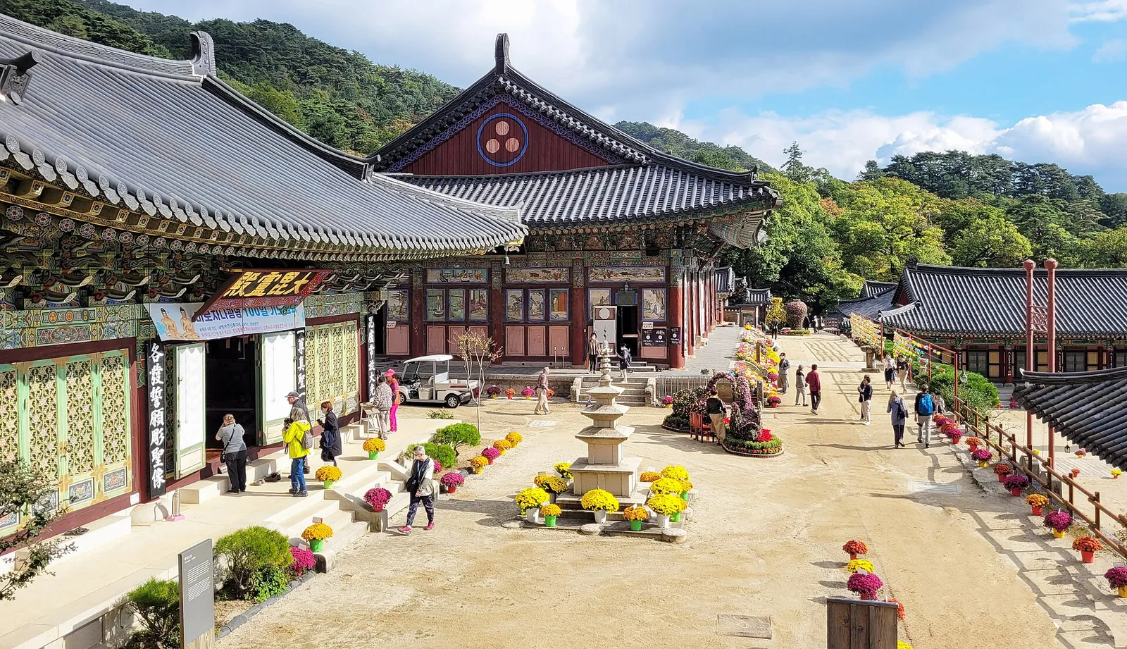 Courtyard of Haeinsa Temple with painted wooden halls, a small stone structure and visitors under a blue sky