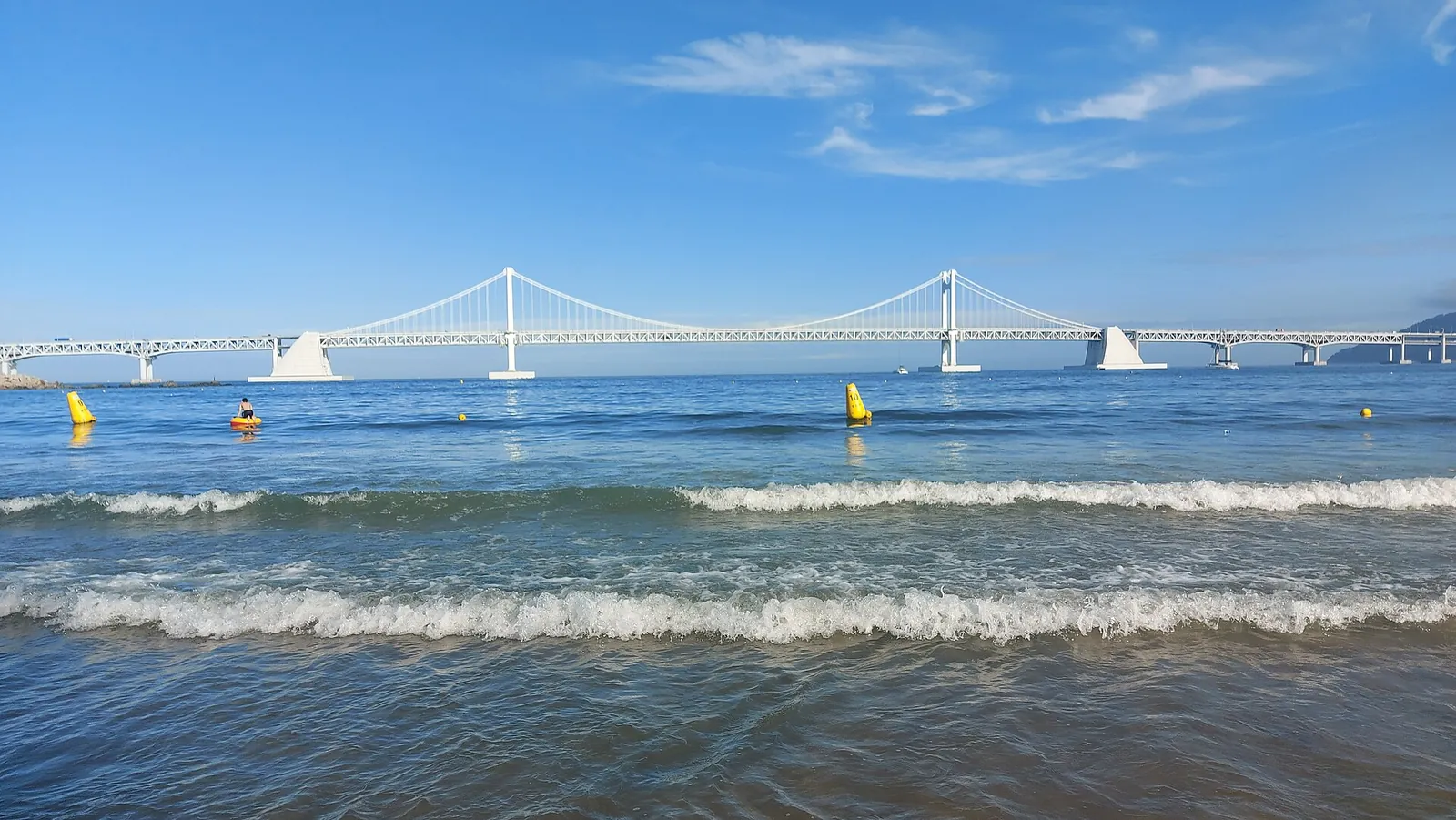 Gwangalli Beach with waves on the sand and Gwangandaegyo suspension bridge across the bay under a blue sky