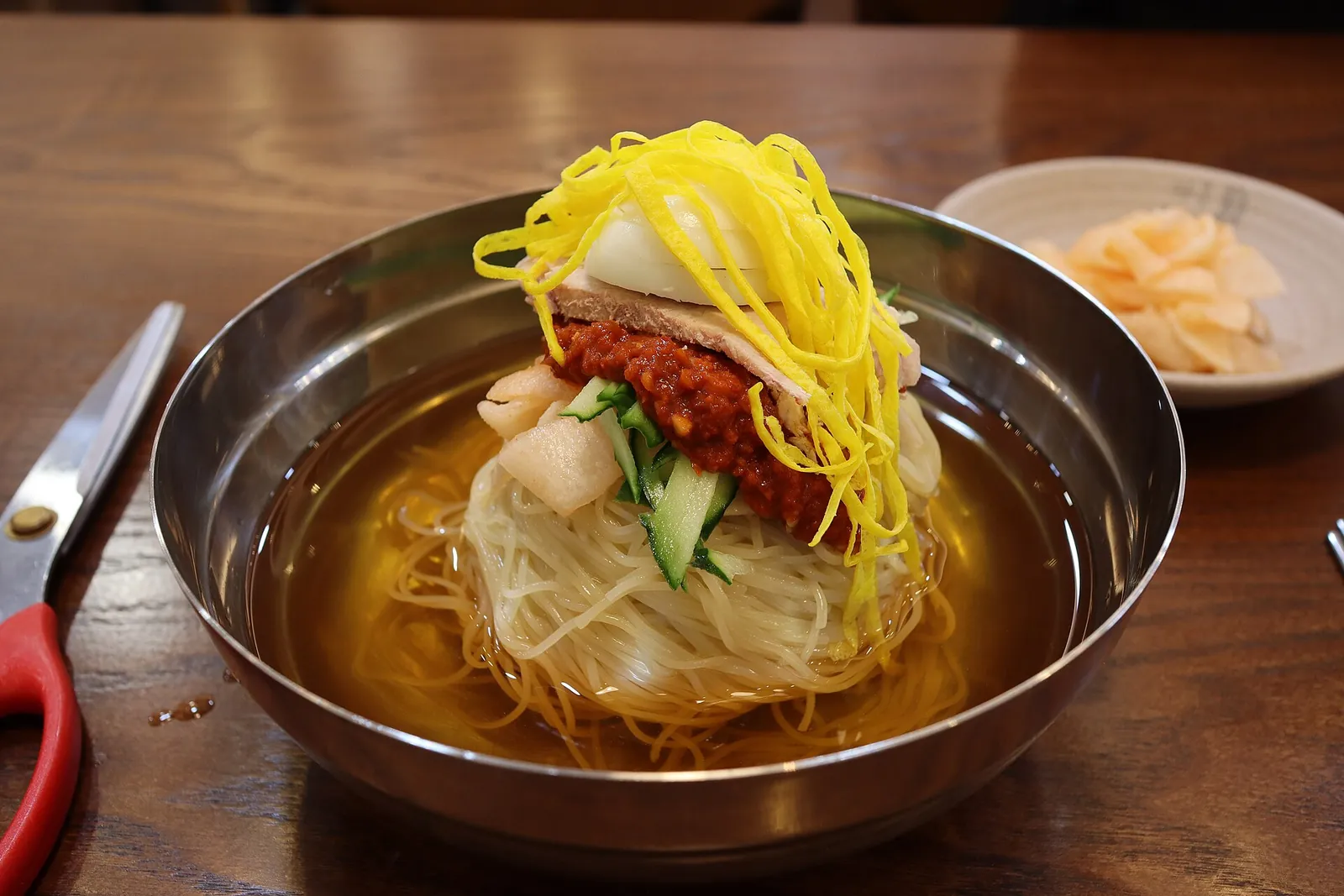 A steel bowl of Busan milmyeon: cold wheat noodles in broth topped with sliced egg, cucumber, meat and red chilli sauce