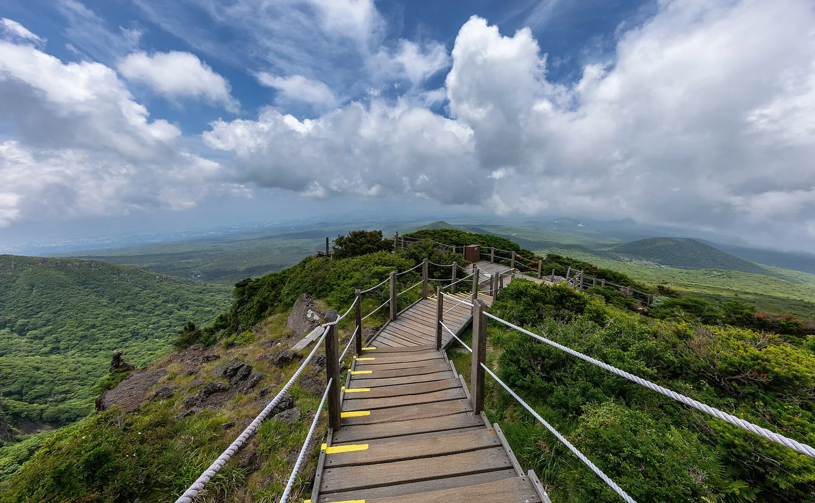 Wooden boardwalk with handrails crossing a green ridge on the Yeongsil Trail under big white clouds