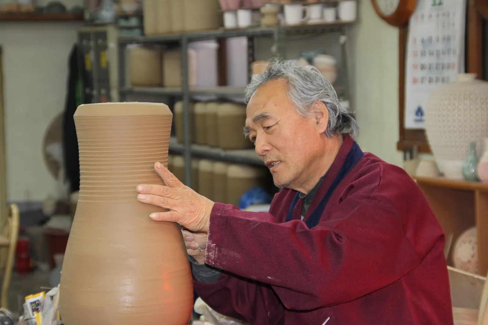 An elderly potter in a red jacket shaping a tall unglazed vase in his Icheon workshop