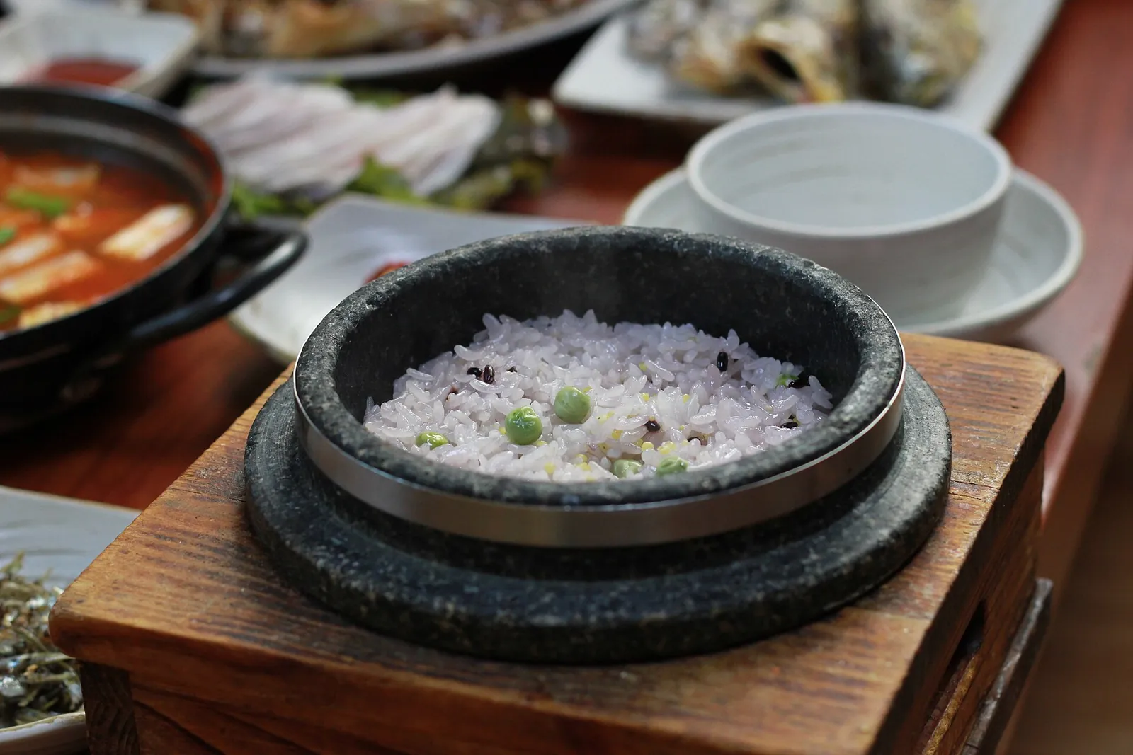 A stone pot of steamed rice with peas and purple rice, surrounded by side dishes and a red stew