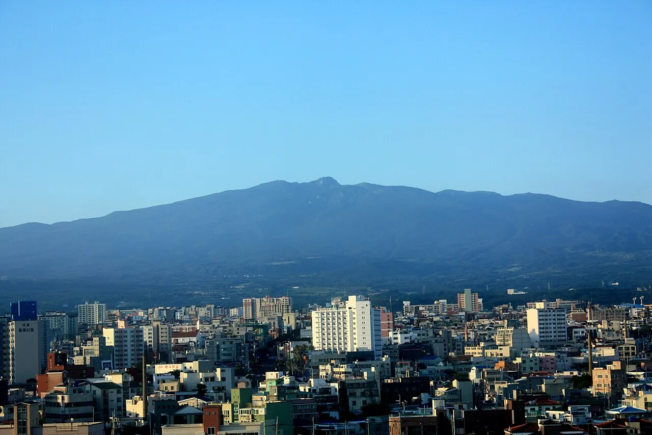 Rooftops and apartment blocks of Jeju City with the long, gentle slope of Hallasan behind under a clear sky