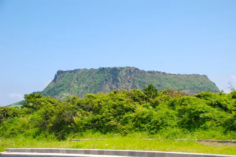 Seongsan Ilchulbong tuff cone rising behind green shrubs under a clear blue sky, Jeju
