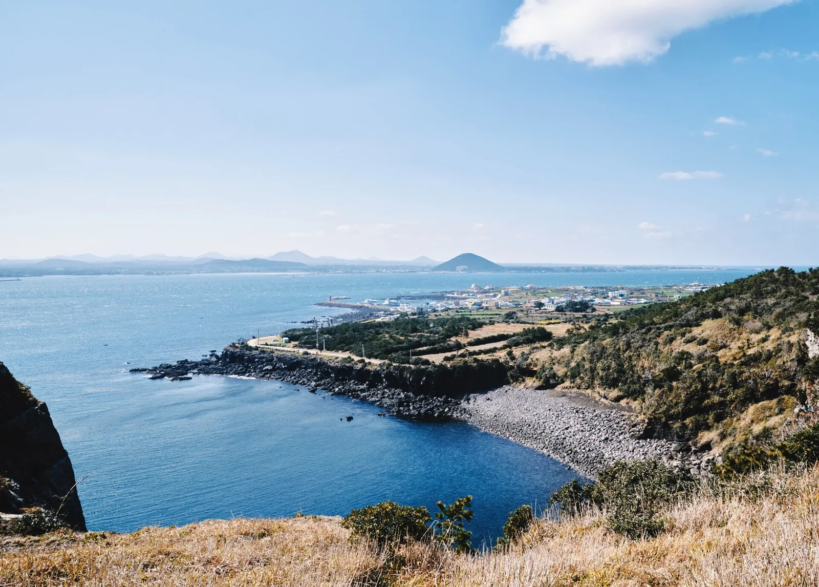 View from Udo Island across blue water and a pebble cove to the Jeju coast and distant volcanic cones