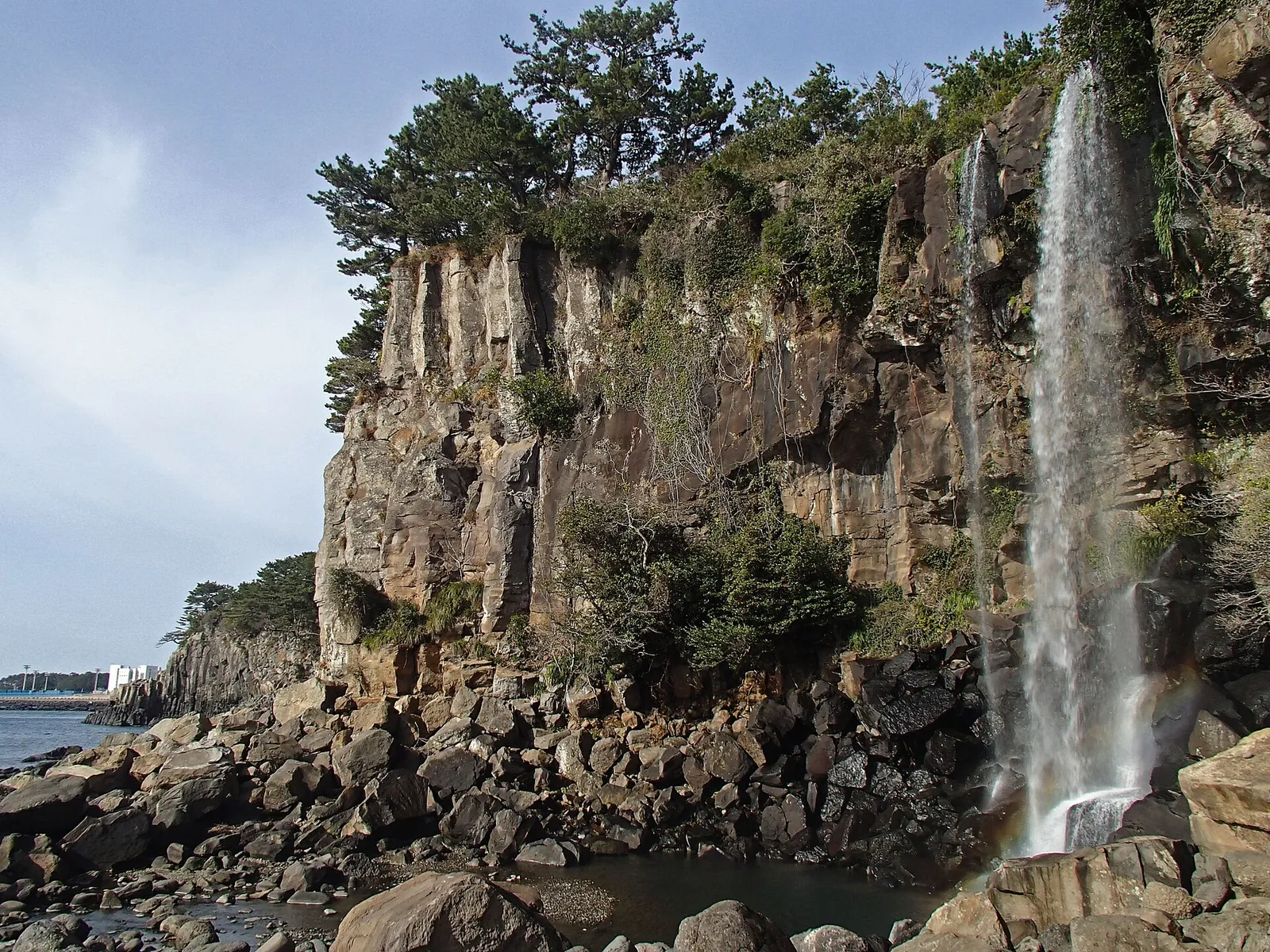 Jeongbang Falls pouring down a dark rock cliff topped with pine trees onto a boulder shore