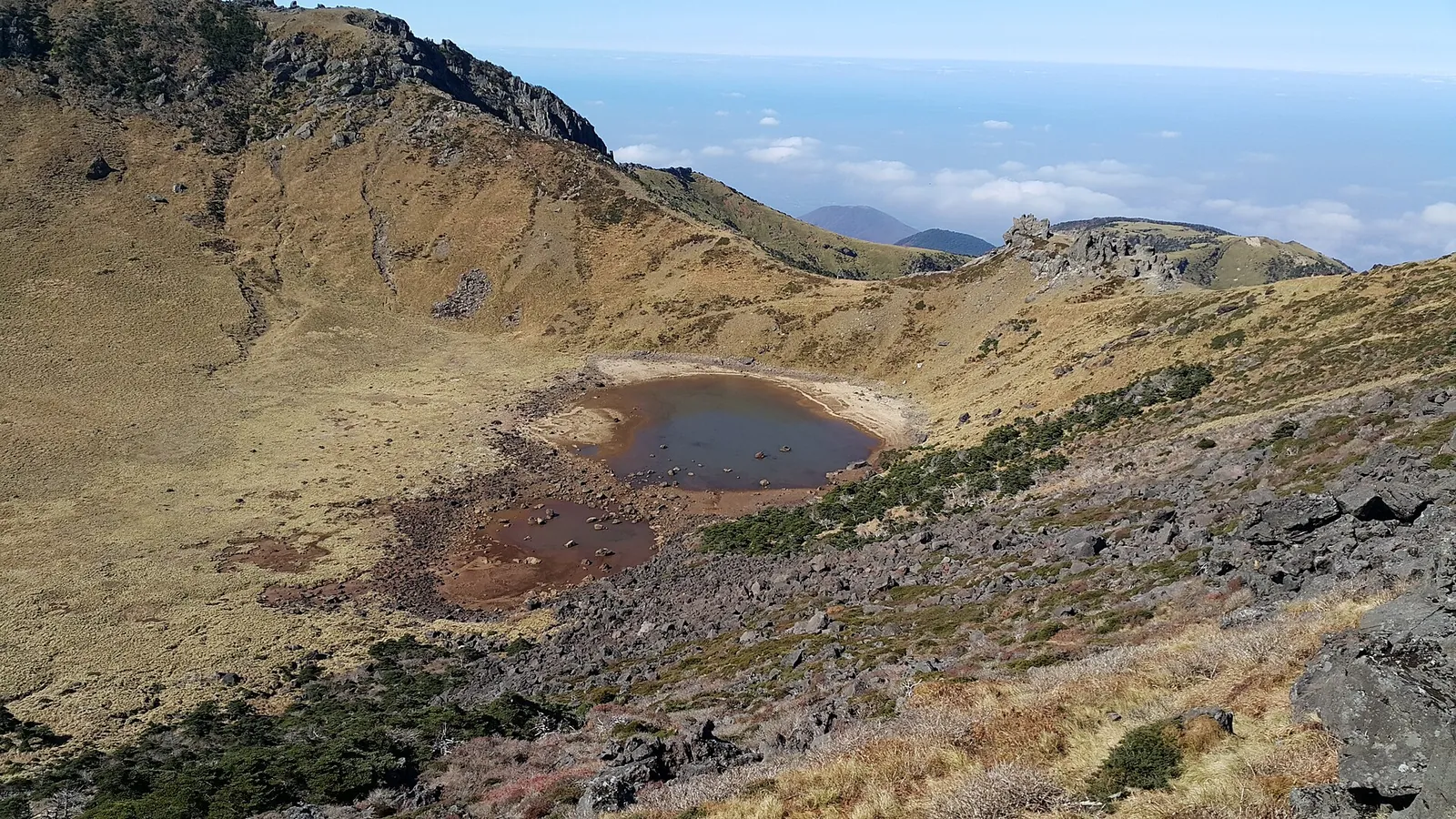 Baengnokdam crater at the summit of Hallasan, a small lake in a brown grassy crater with sea and clouds beyond