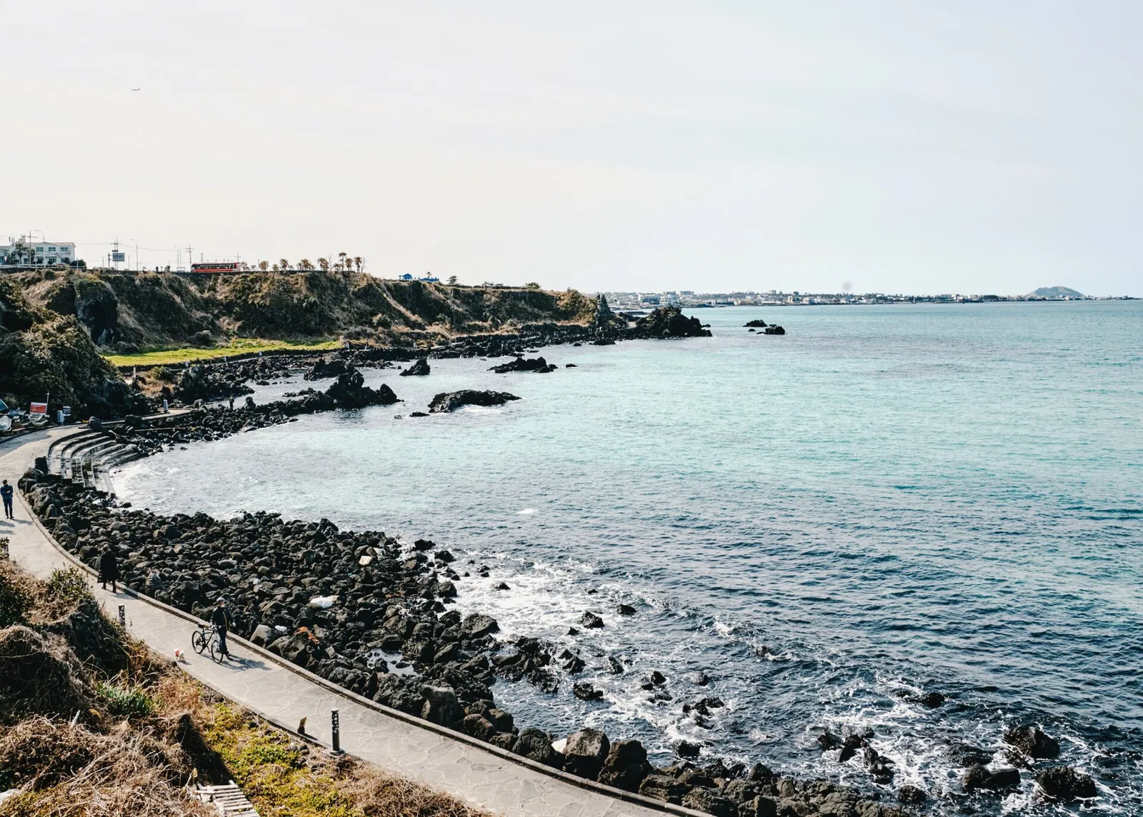 A paved coastal path curving along black volcanic rocks and blue water on Olle Route 15-B in Aewol