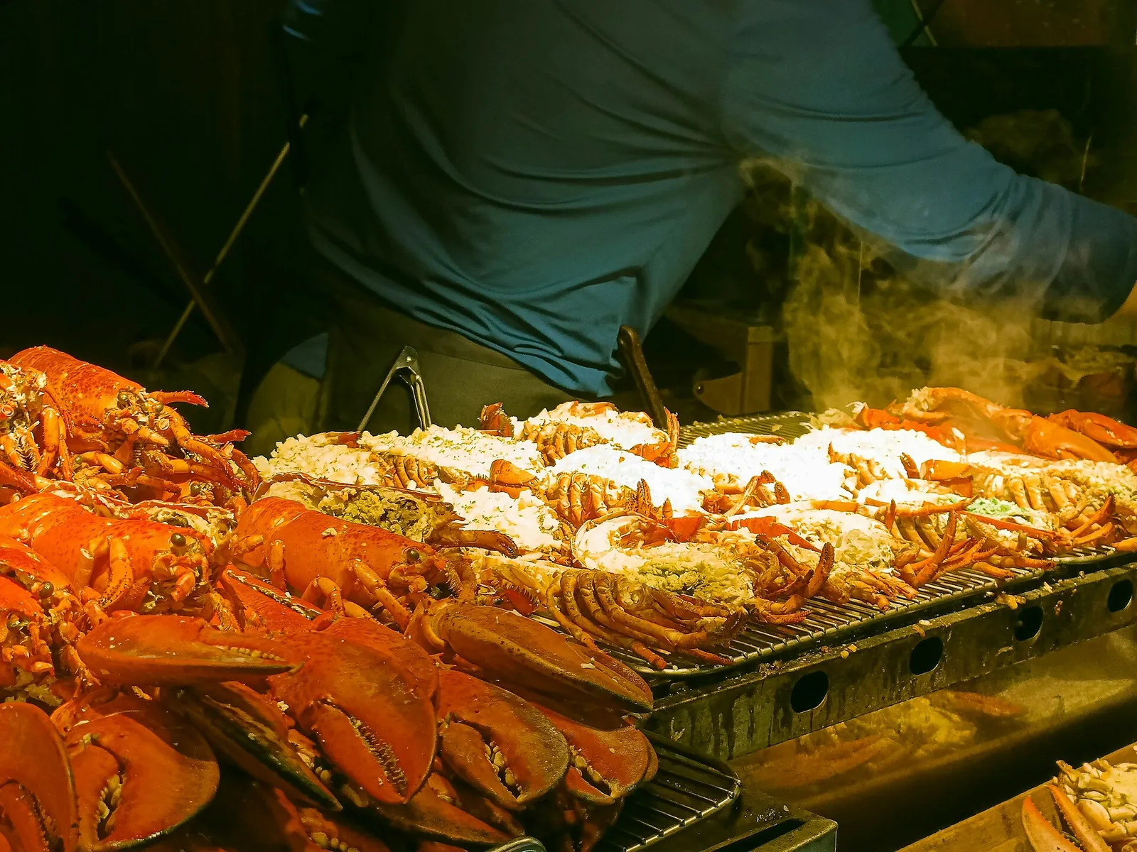 Halved lobsters with a white topping on a steaming grill beside a pile of whole cooked lobsters and crabs at a Dongmun Market stall, a vendor in a blue shirt behind