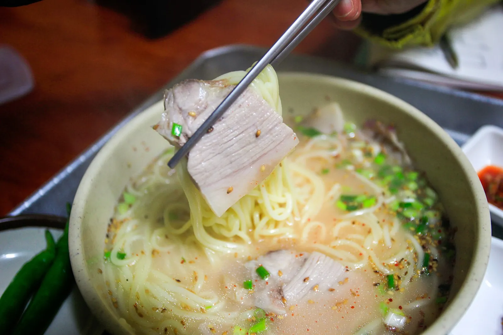 A bowl of gogi-guksu, noodles in a pale pork broth with sliced pork and green onion, lifted with chopsticks
