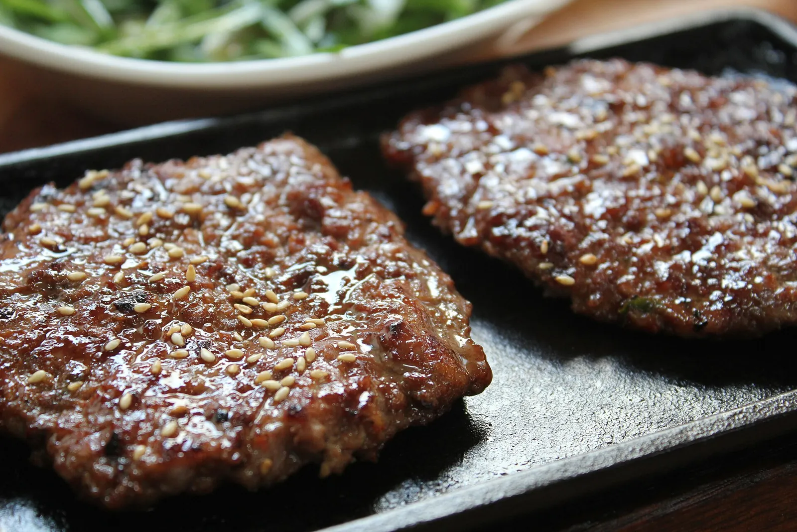 Two grilled tteokgalbi patties glazed and scattered with sesame on a hot plate