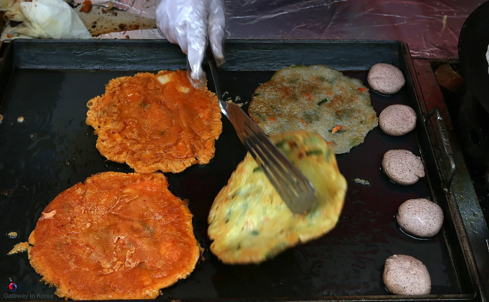 Red and green vegetable pancakes and small round pancakes frying on a flat griddle at a market stall