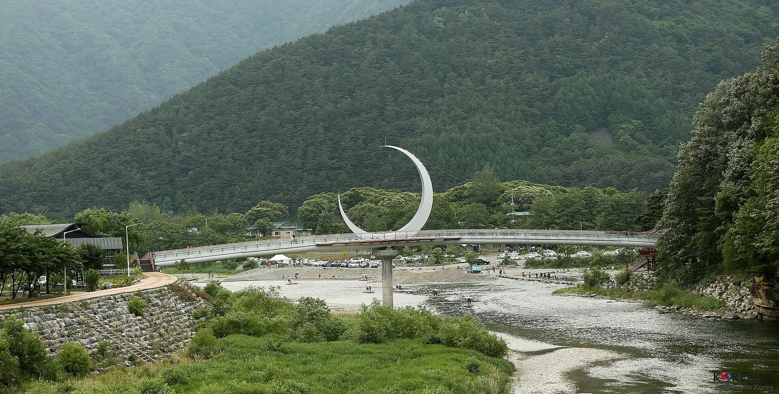 A wide shallow river with a crescent-moon sculpture on the bank and a bridge, surrounded by green mountains in Jeongseon County