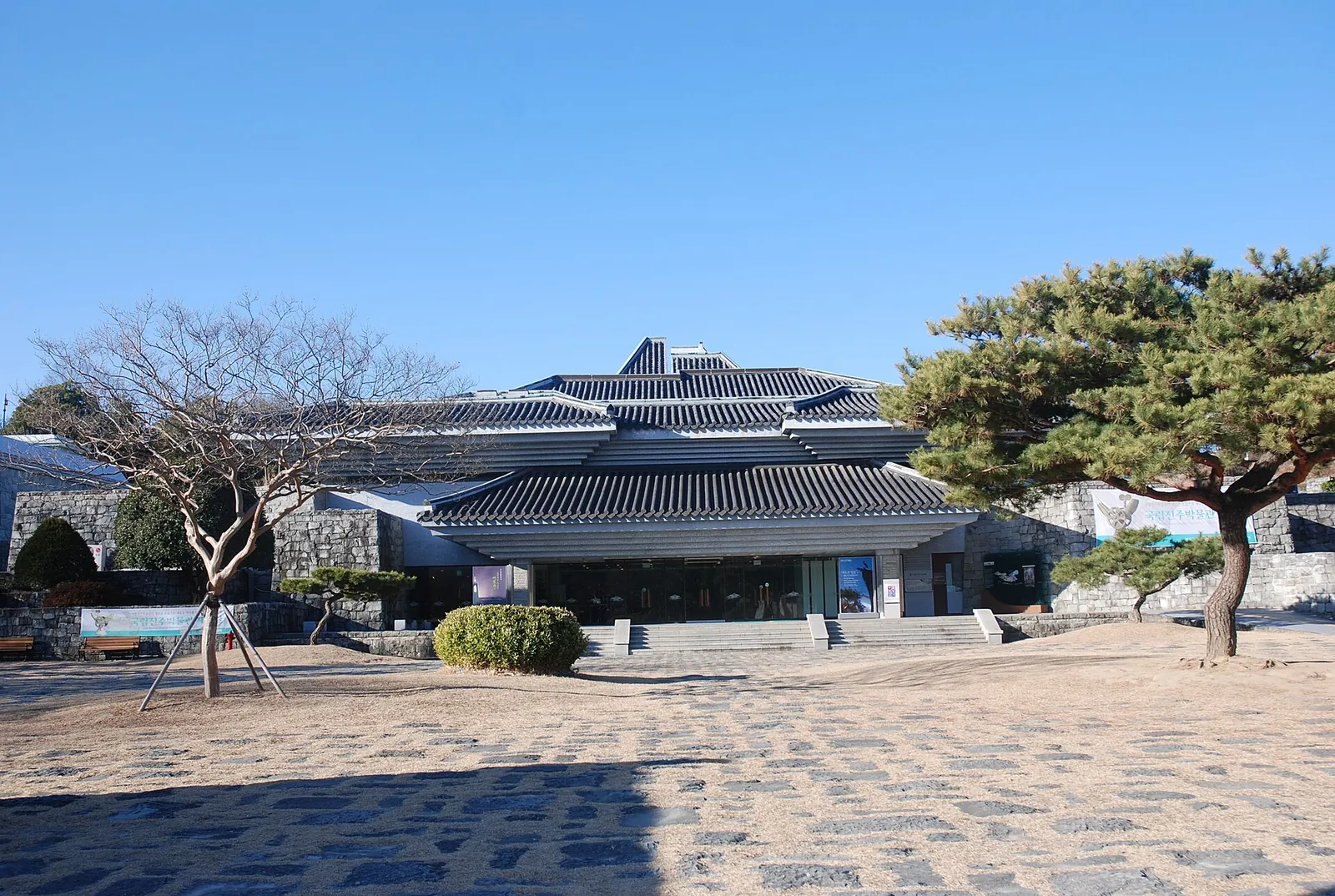 Low tiled-roof building of Jinju National Museum behind a paved courtyard and pine trees inside Jinjuseong Fortress