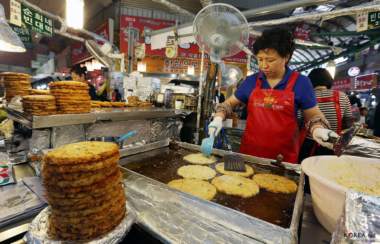 A cook frying mung bean pancakes on a griddle beside a stack of finished bindaetteok at Gwangjang Market