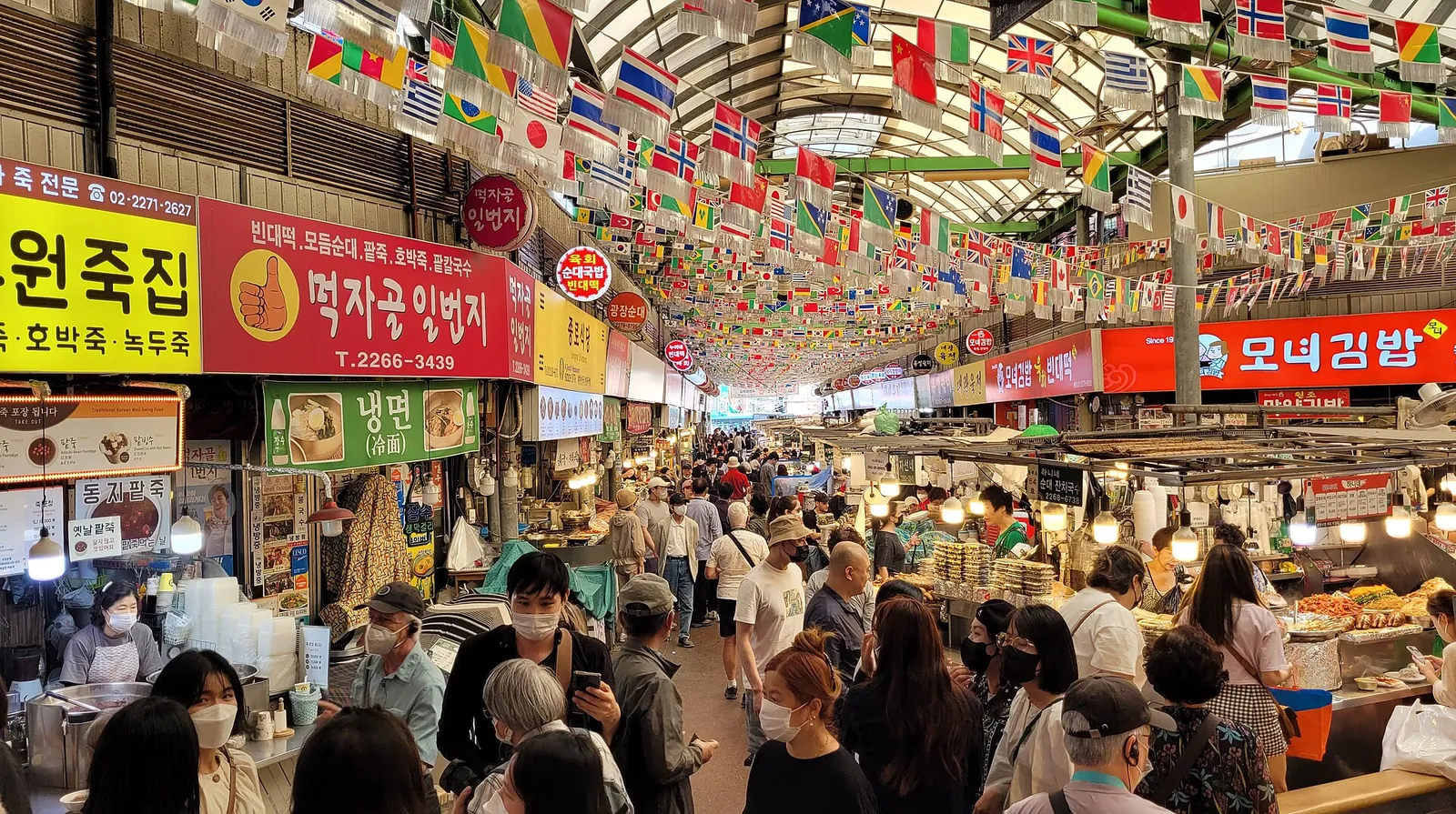 Crowded central aisle of Gwangjang Market in Seoul under colourful flags, with food stalls and shop signs on both sides