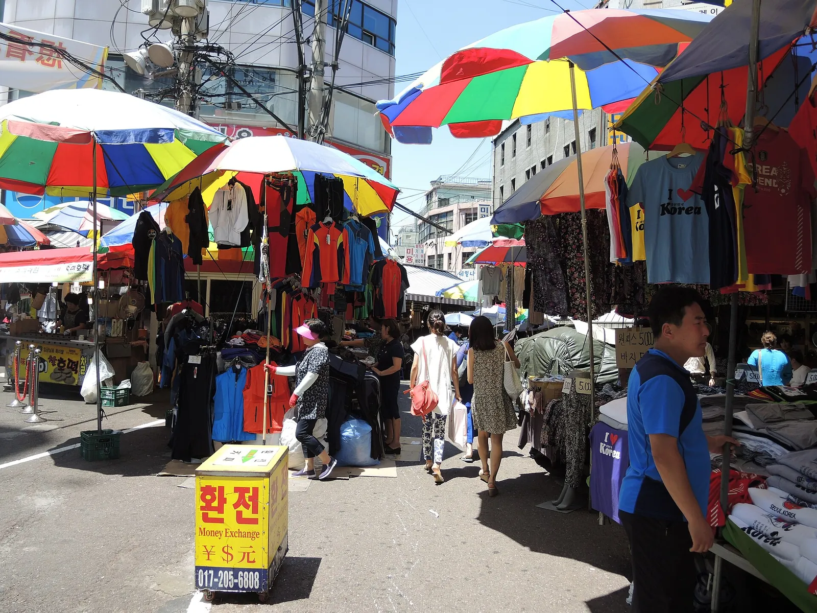 Clothing stalls under striped umbrellas at Namdaemun Market in Seoul, with a yellow currency exchange sign in the foreground