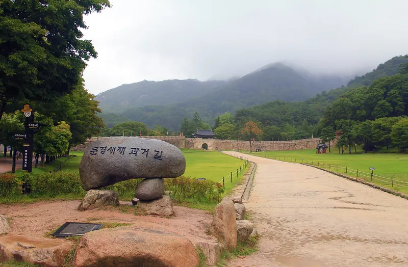 Juheulgwan, the first gate of Mungyeong Saejae, at the end of a path across green lawns, with a carved stone reading Mungyeong Saejae Gwageo-gil in the foreground
