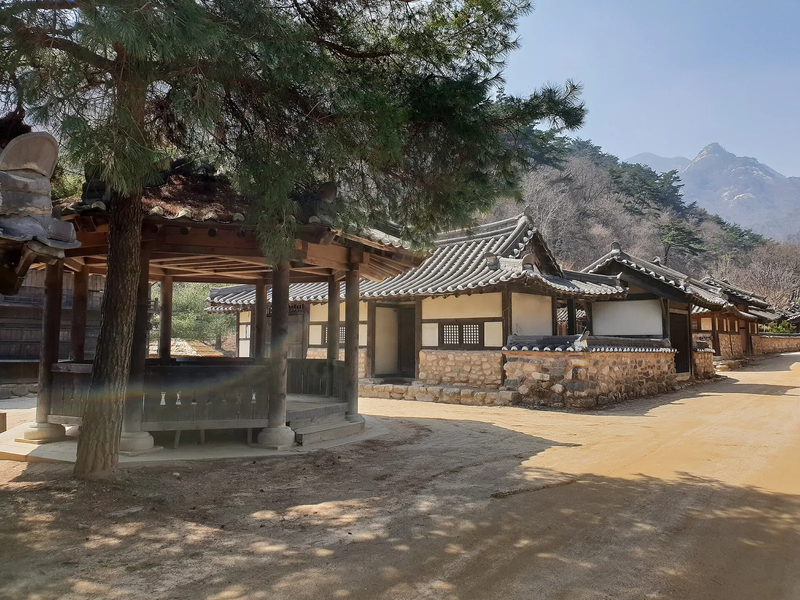 Tiled hanok buildings and a wooden pavilion on a dirt lane at the Mungyeongsaejae Open Set, with mountains behind