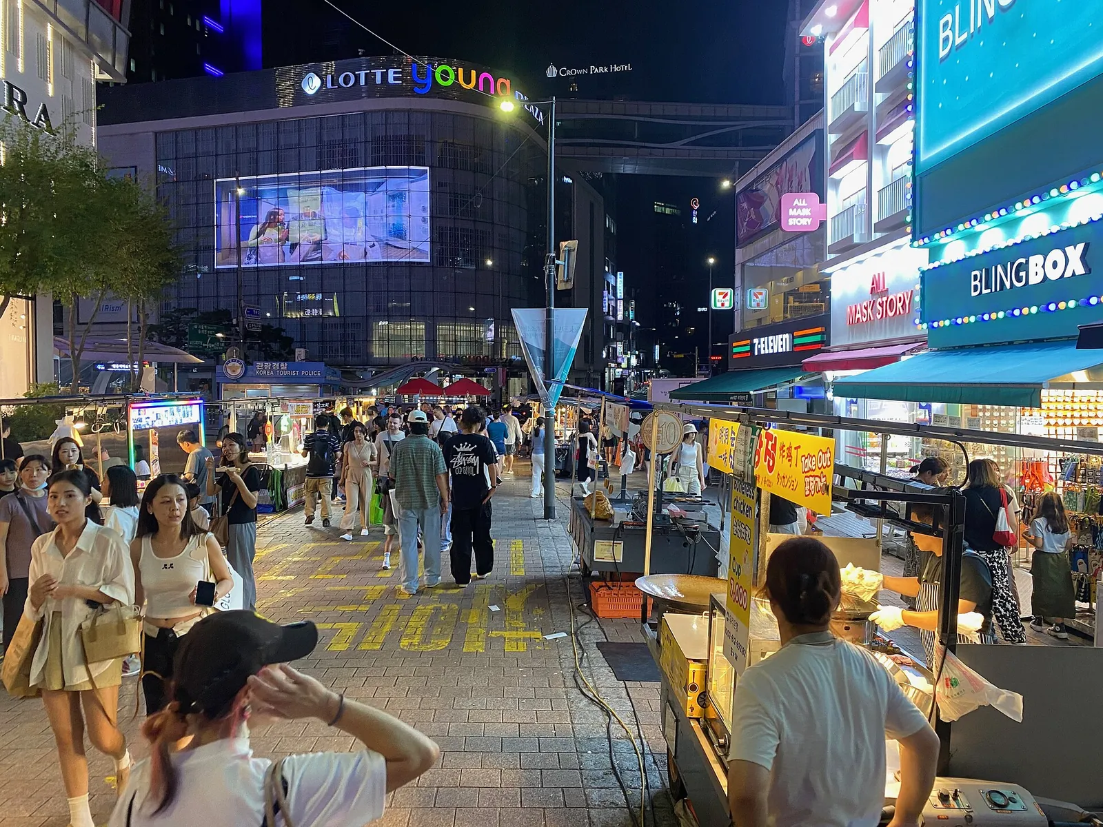 Crowds walking past lit street-food stalls on a Myeongdong shopping street at night, with department-store signs behind