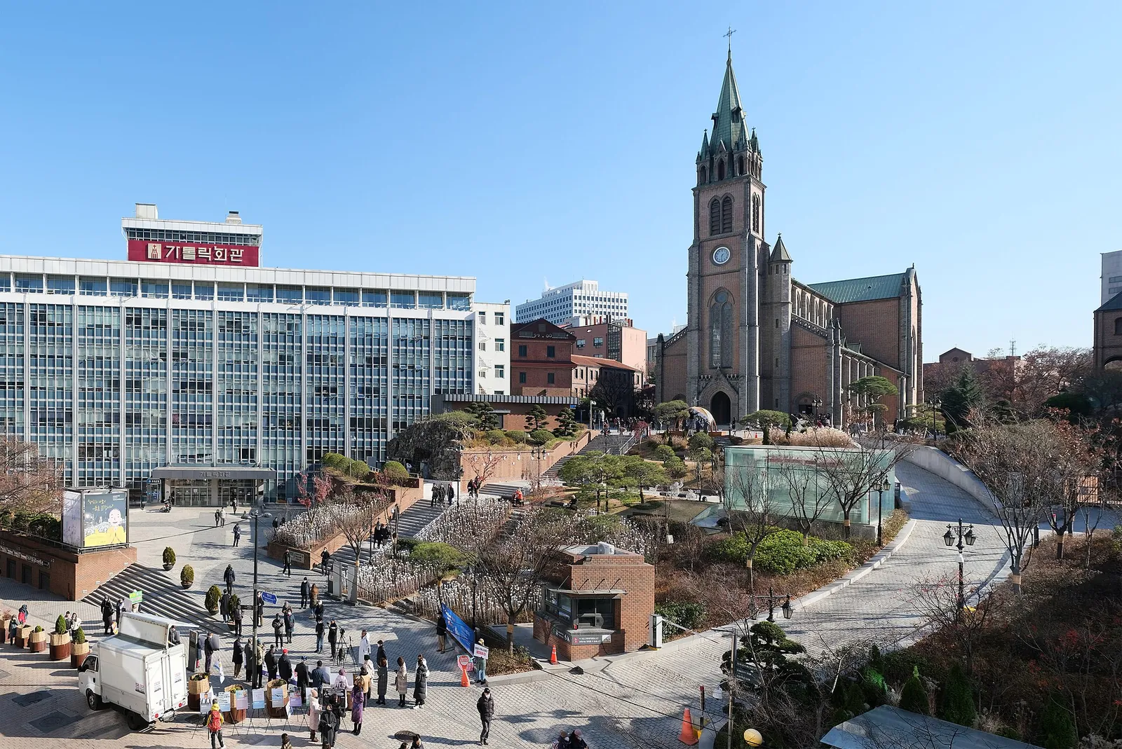 Red-brick Gothic spire of Myeongdong Cathedral above its terraced forecourt and surrounding office buildings