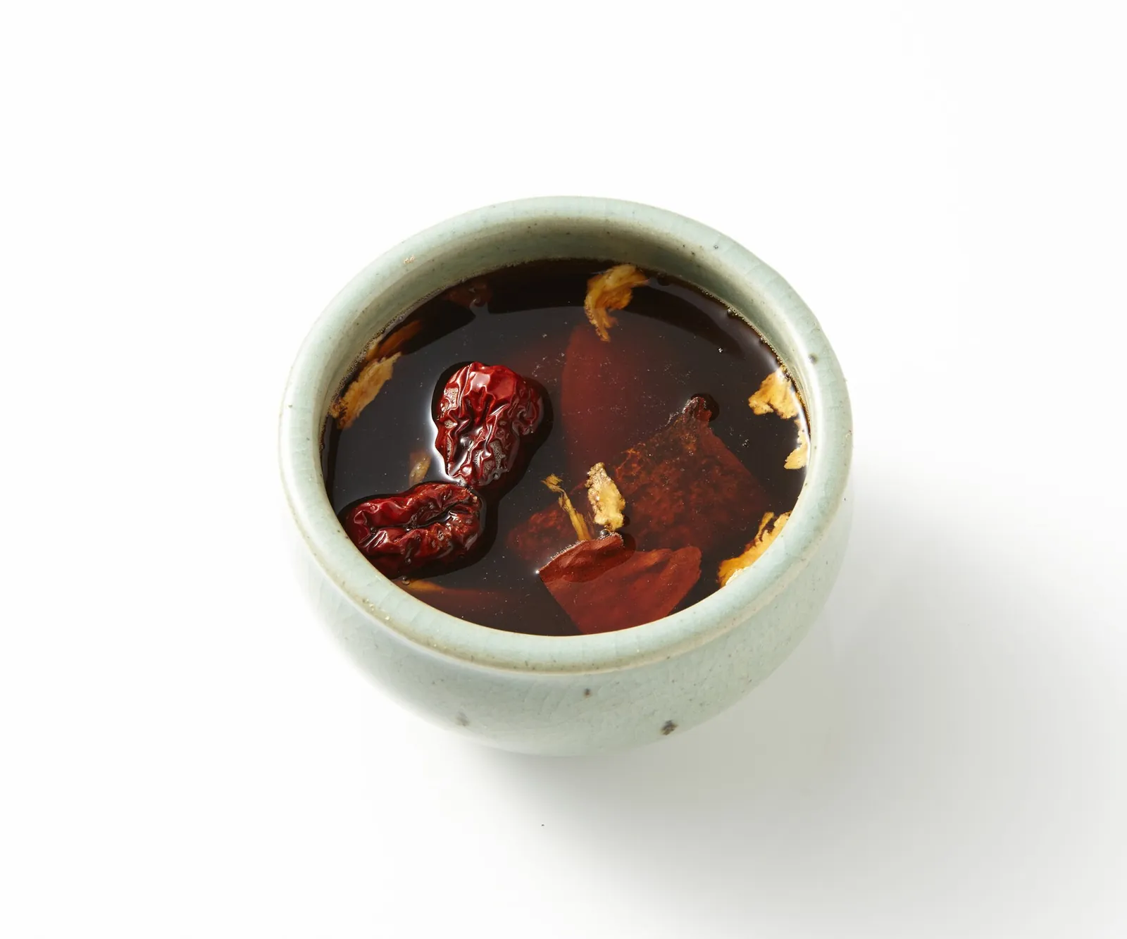A small celadon-glazed cup of dark ssanghwatang herbal tea with dried jujubes and pieces of herbs floating in it