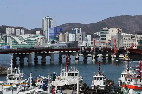 Yeongdo Bridge s červeným zábradlím přes přístav za kotvícími rybářskými loděmi, s budovou Jagalchi Market s vlnovitou střechou vlevo