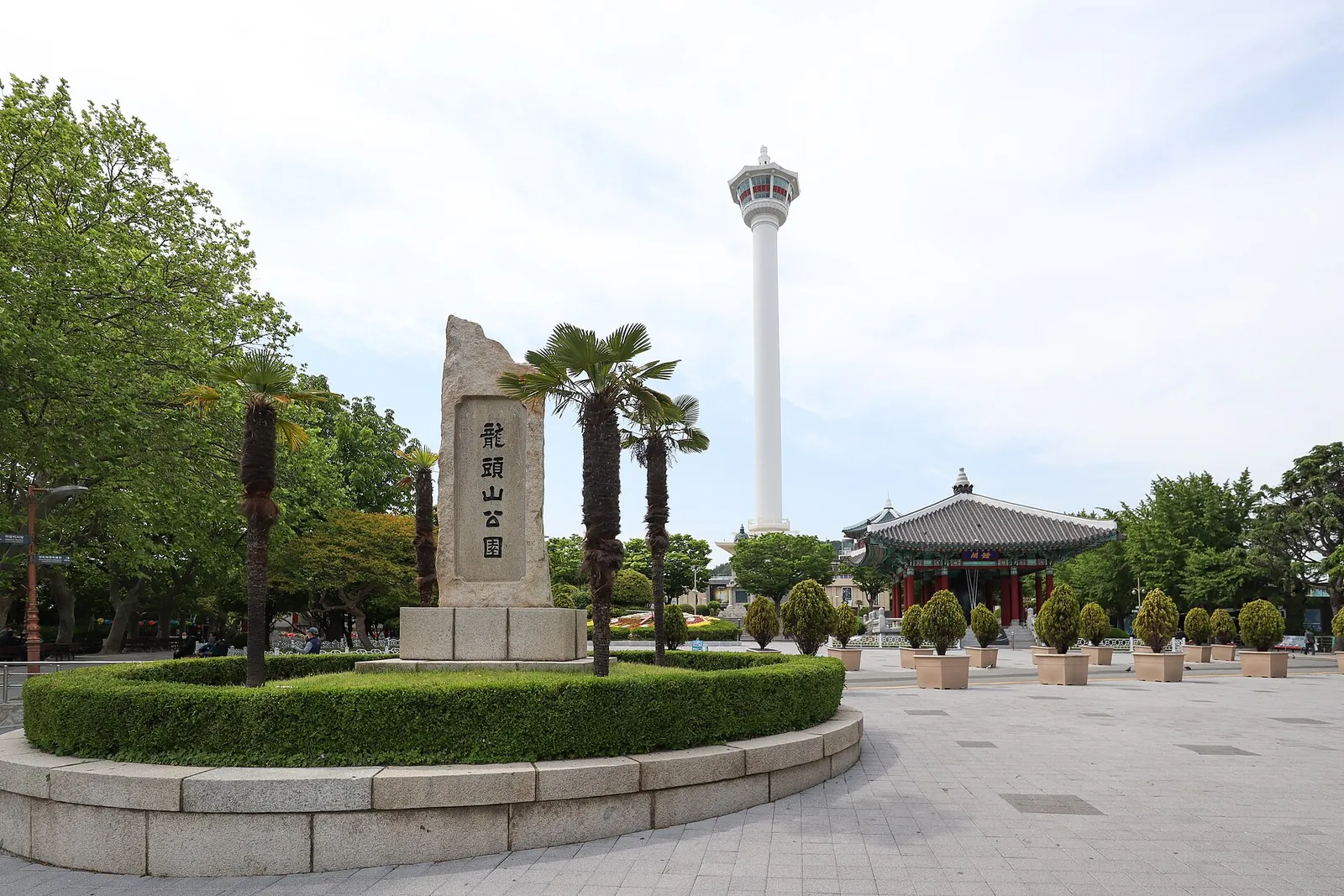 Busan Tower rising above the trees and memorial stone of Yongdusan Park on a sunny day