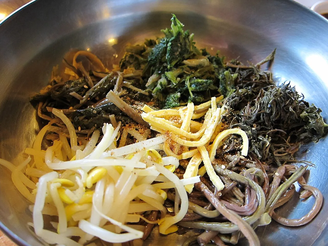 A metal bowl of sanchae bibimbap with mountain greens, fernbrake, bean sprouts and strips of egg over rice