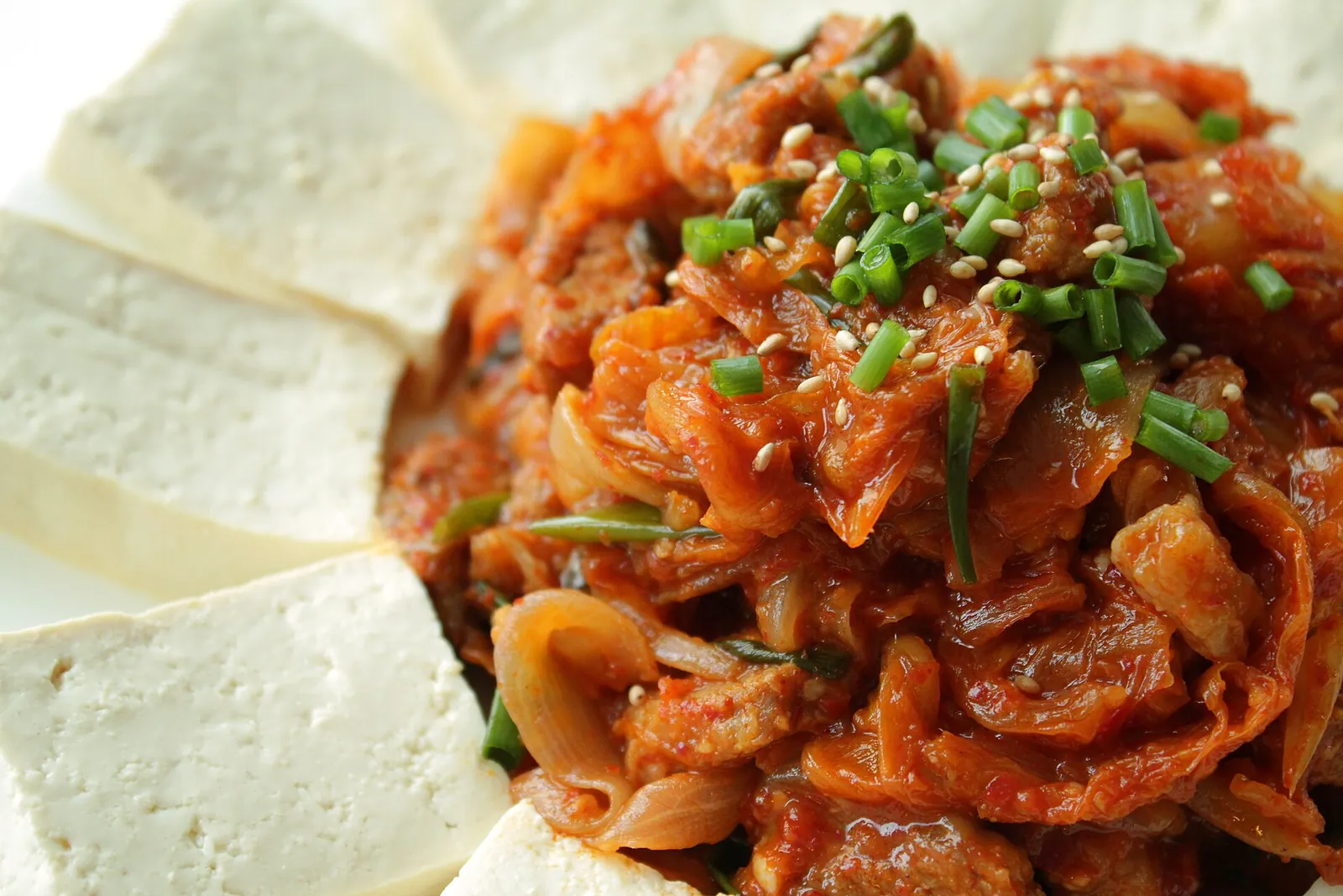 Plate of warm tofu slices around stir-fried kimchi and pork topped with spring onion and sesame
