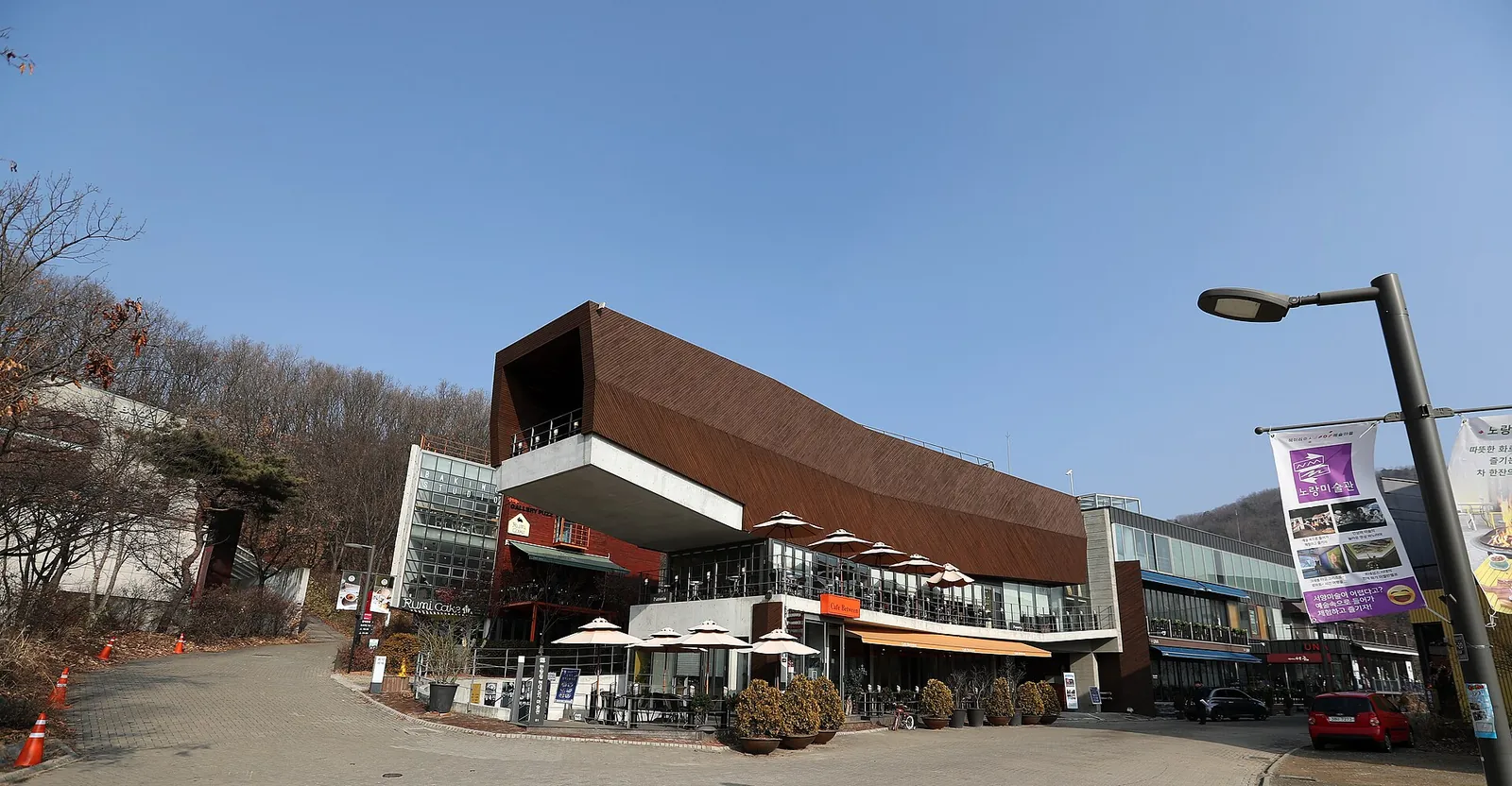 Rust-coloured modern gallery building and cafés along a street in Heyri Art Valley under a blue sky