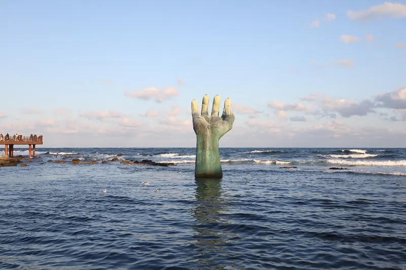 The bronze Hand of Harmony sculpture rising from the sea at Homigot, with a pier on the left and a bright sky
