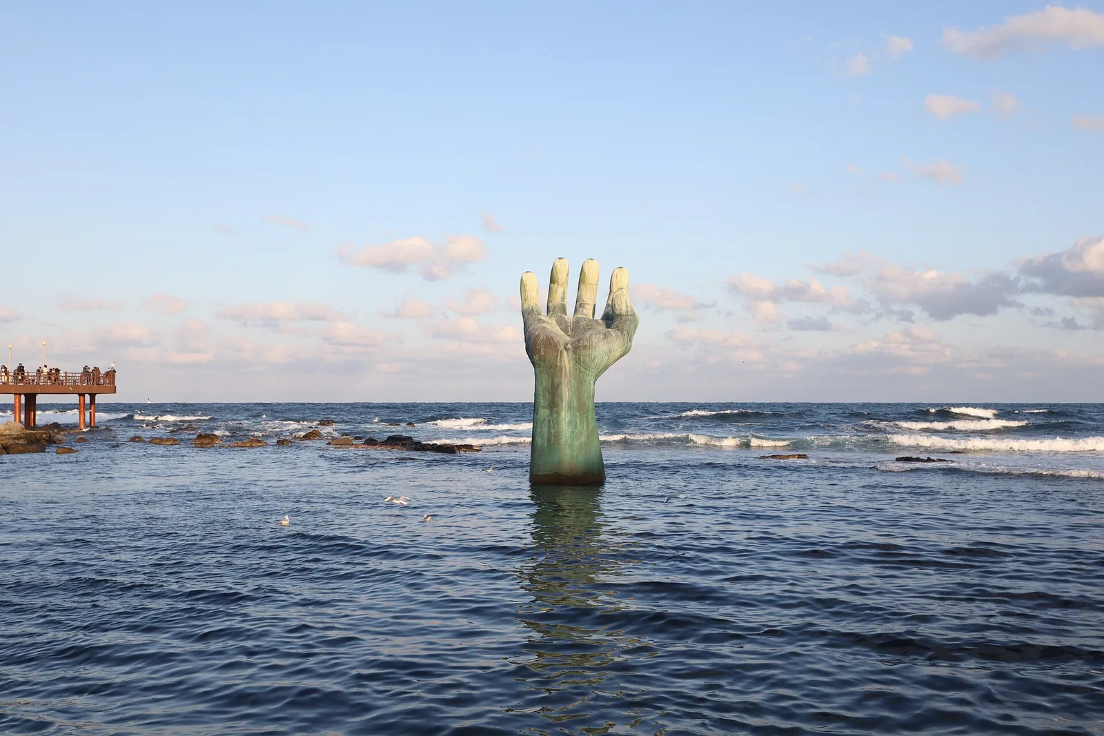 The bronze Hand of Harmony sculpture rising from the sea at Homigot, with a pier on the left and a bright sky