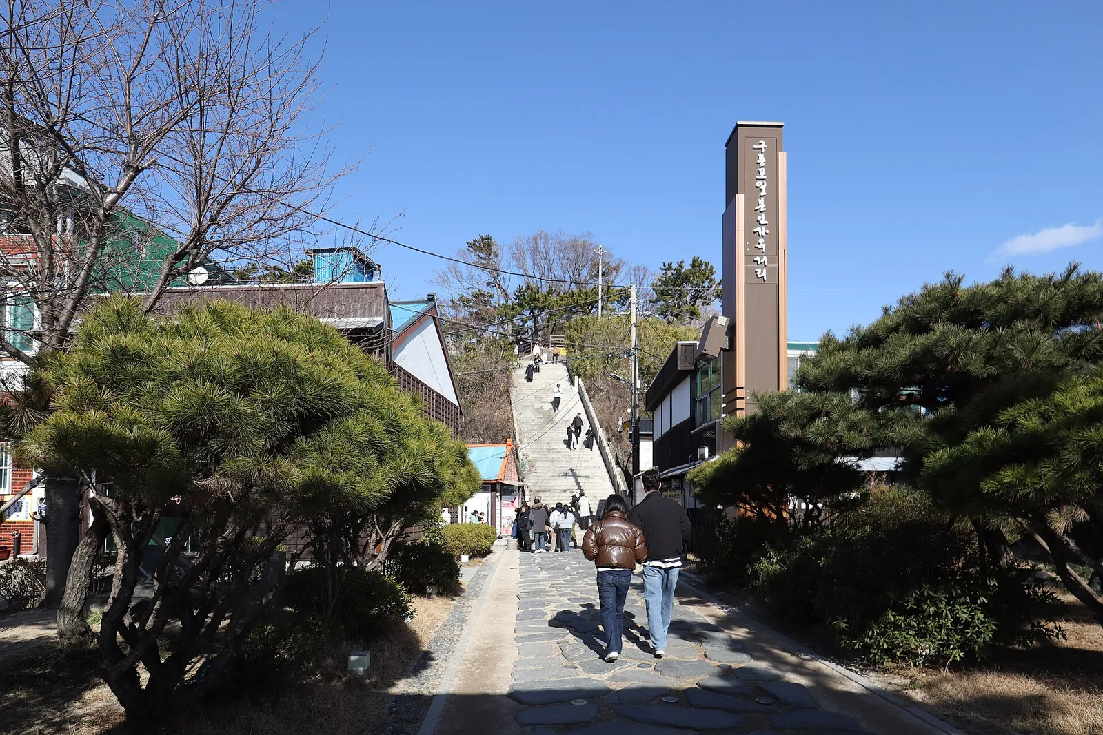 A sloping street in Guryongpo with pine trees, a stone staircase climbing to the park and a tall sign at the top