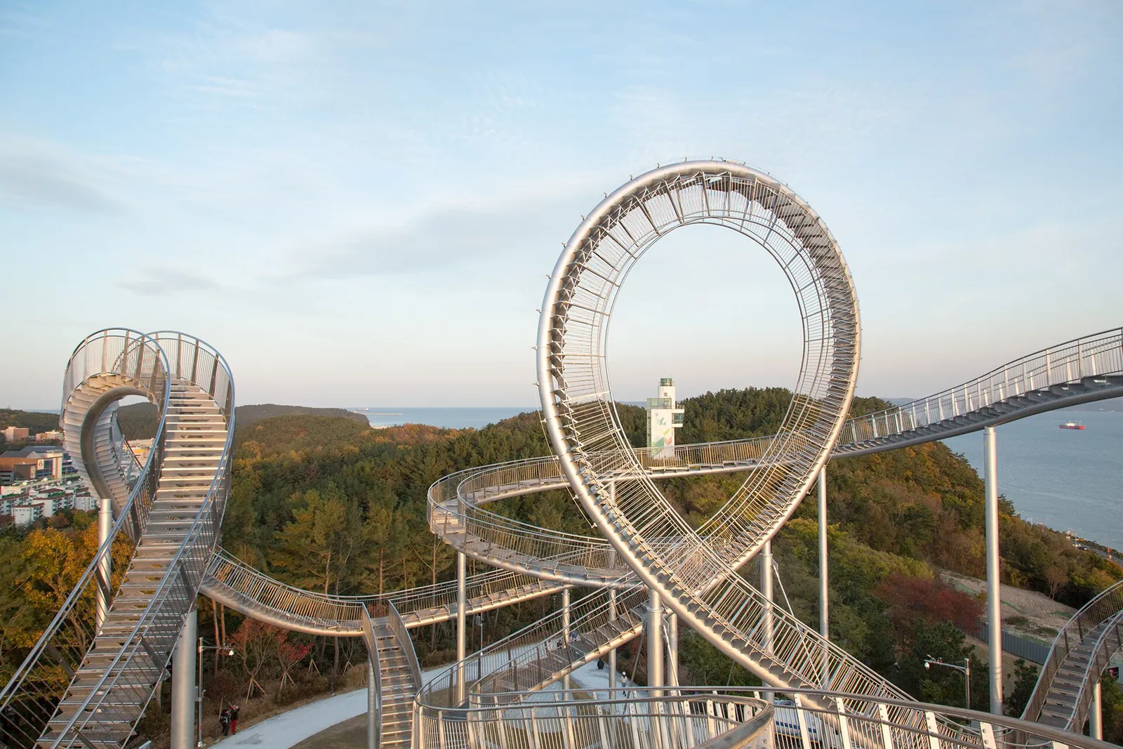 The looping steel stairway of Space Walk above the trees of Hwanho Park, with the sea behind