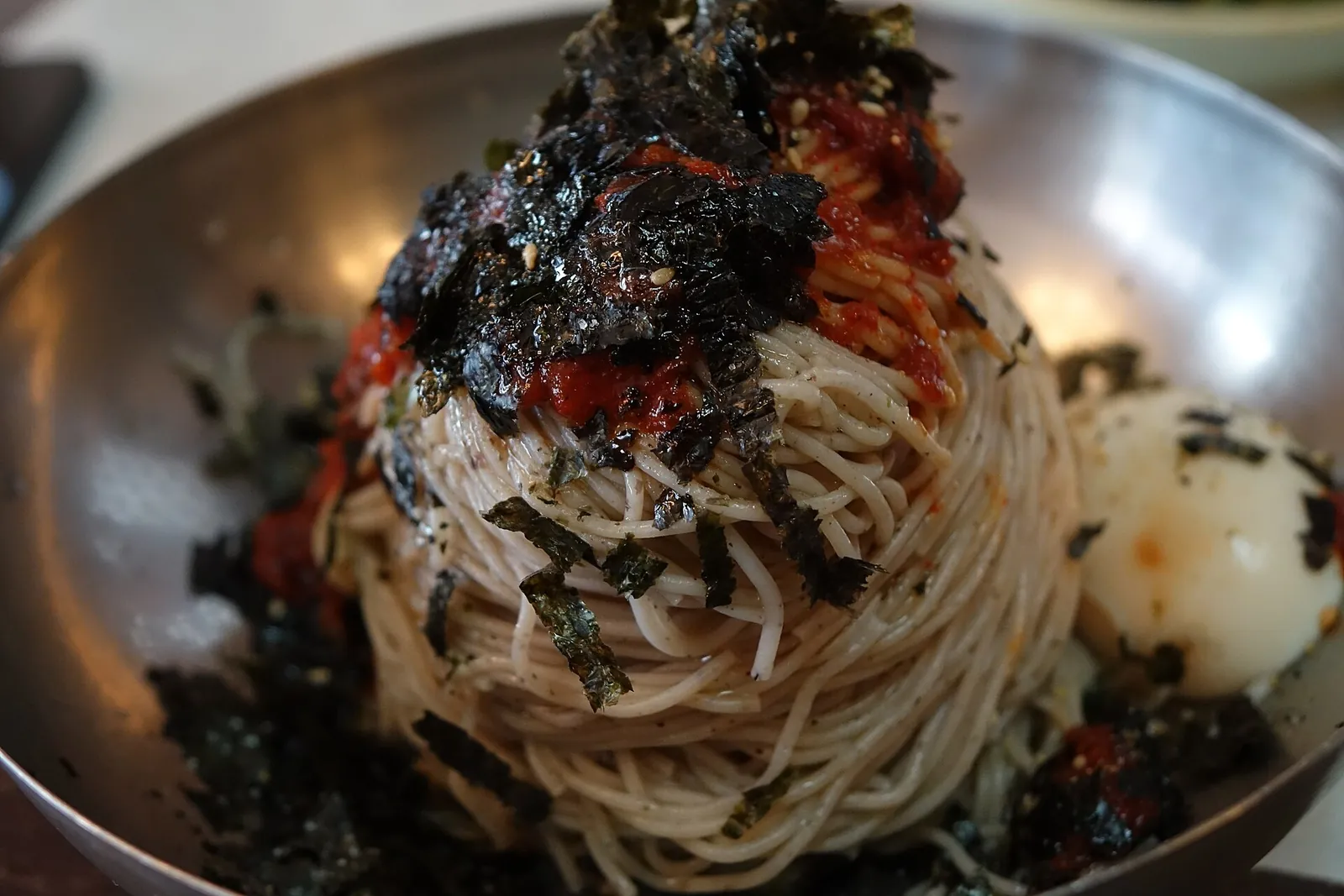 A steel bowl of spicy buckwheat noodles topped with red chilli sauce, shredded laver and half a boiled egg