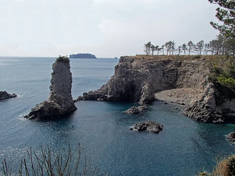 The Oedolgae sea stack rising from calm blue water beside a pine-topped cliff, with an island on the horizon