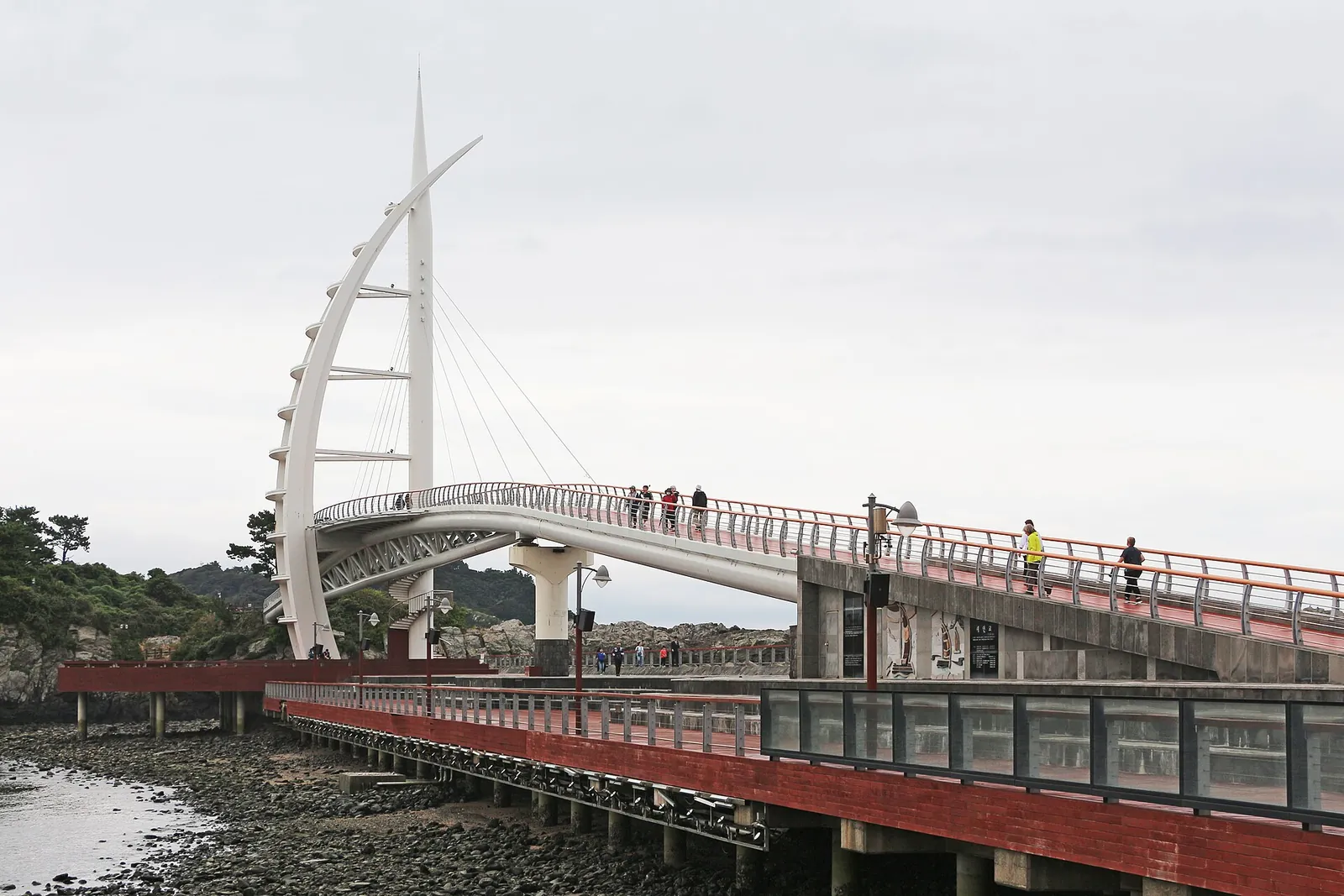 The white sail-shaped pylon of Saeyeongyo Bridge above a red-railed walkway at Seogwipo harbour