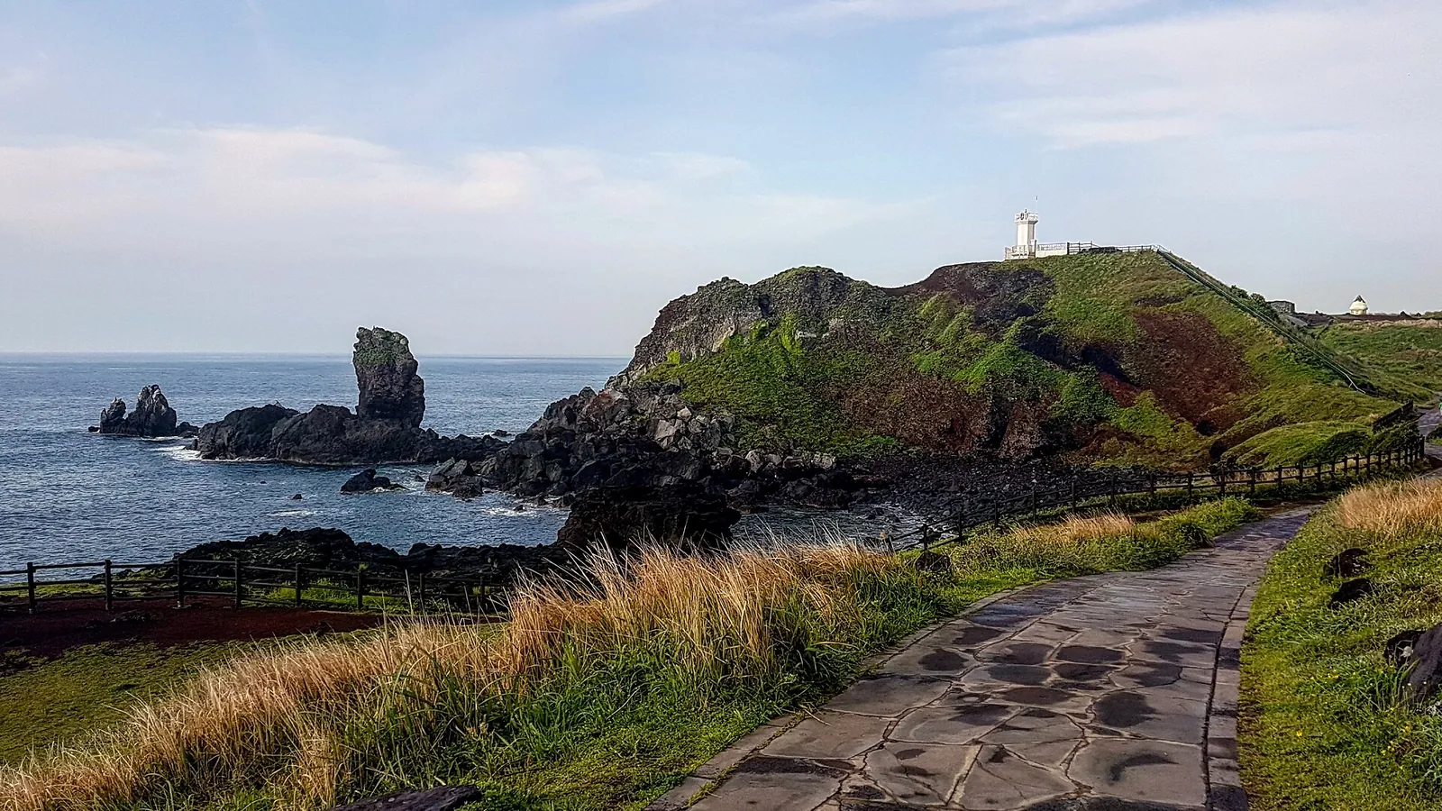 Coastal path through dry grass at Seopjikoji with basalt sea stacks and a white lighthouse on a red-soiled hill