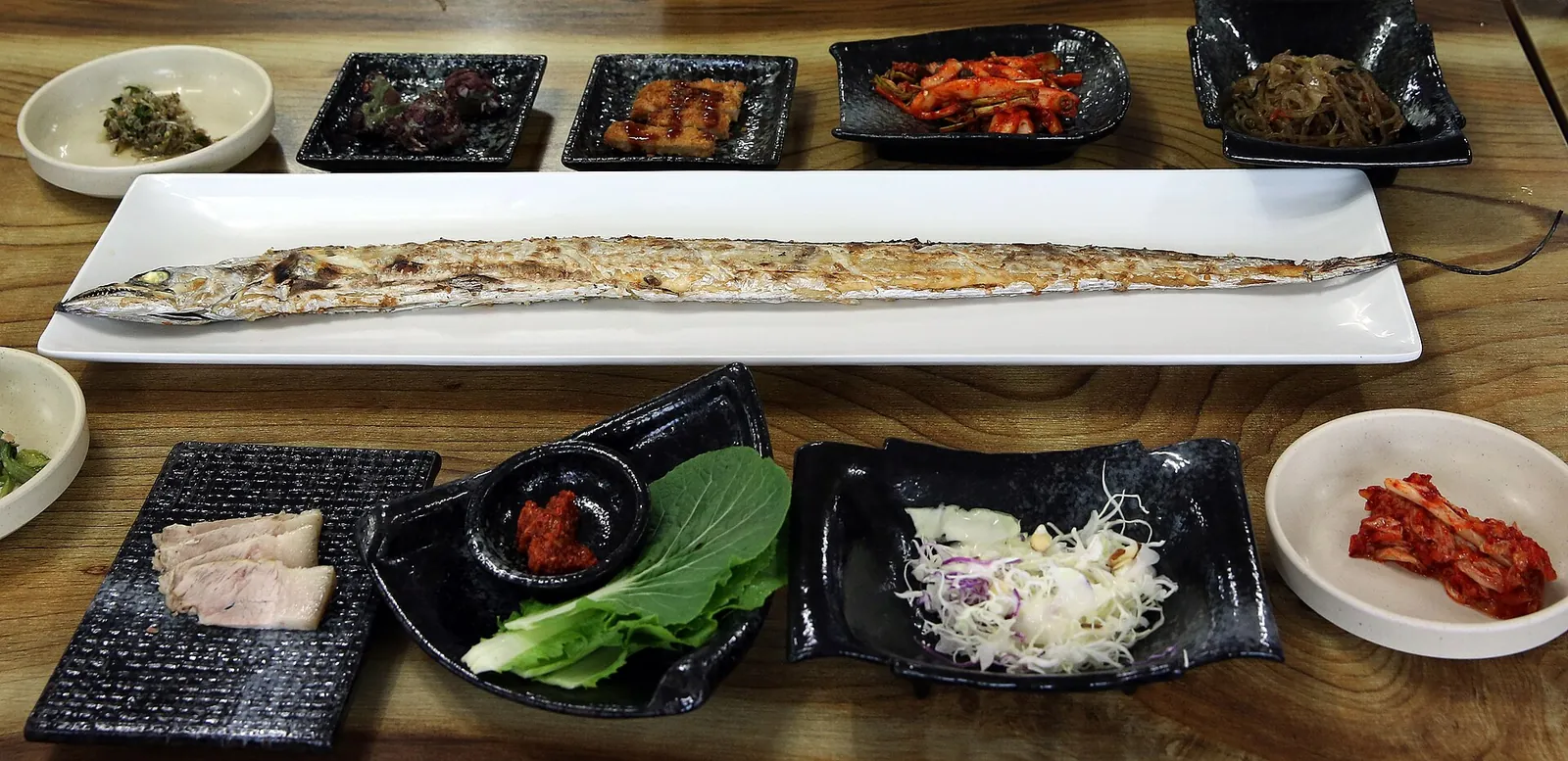 A whole grilled cutlassfish on a long white platter surrounded by small Korean side dishes