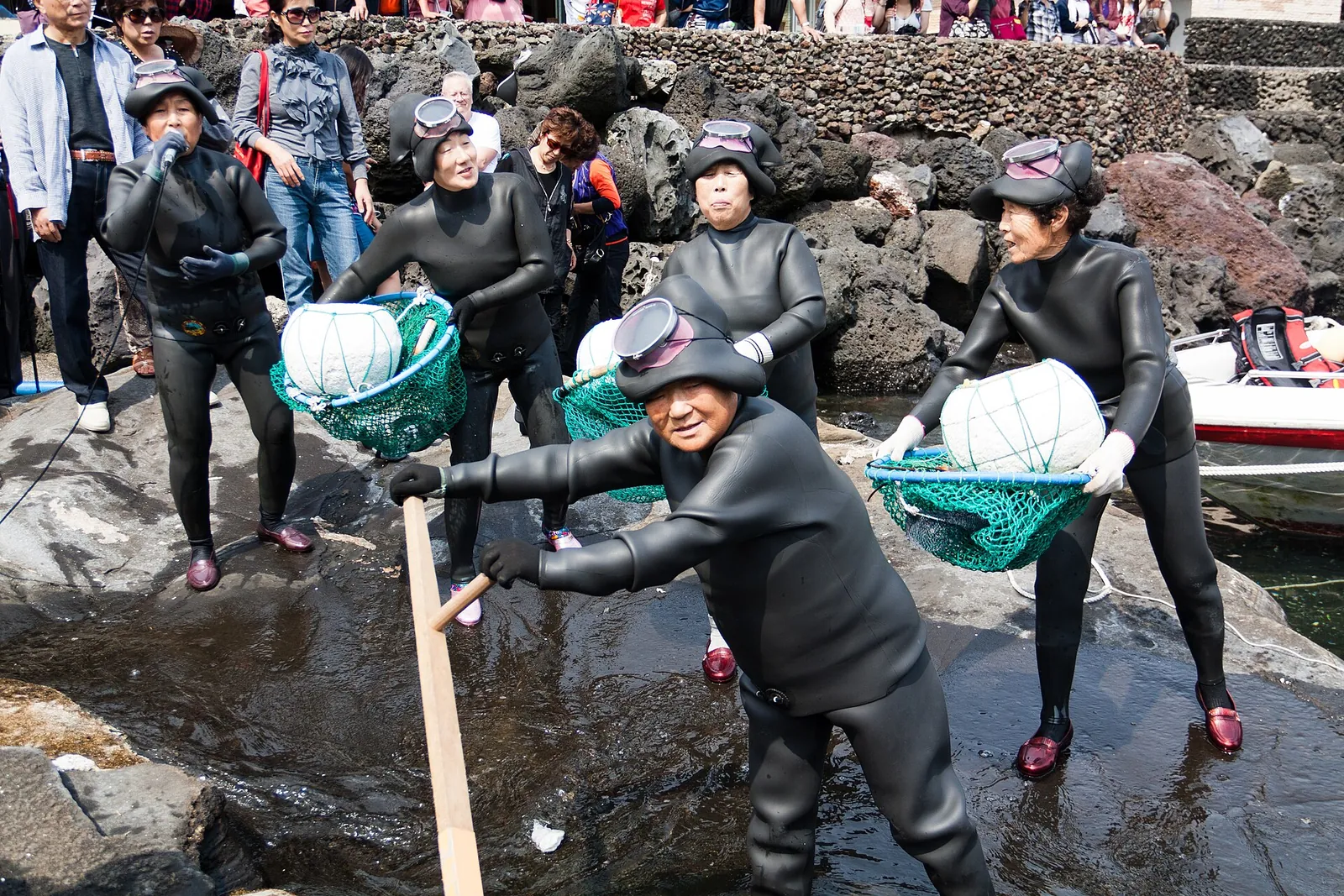 Jeju haenyeo in black wetsuits and diving masks carrying nets with white floats up a rocky shore, one speaking into a microphone, watched by a crowd