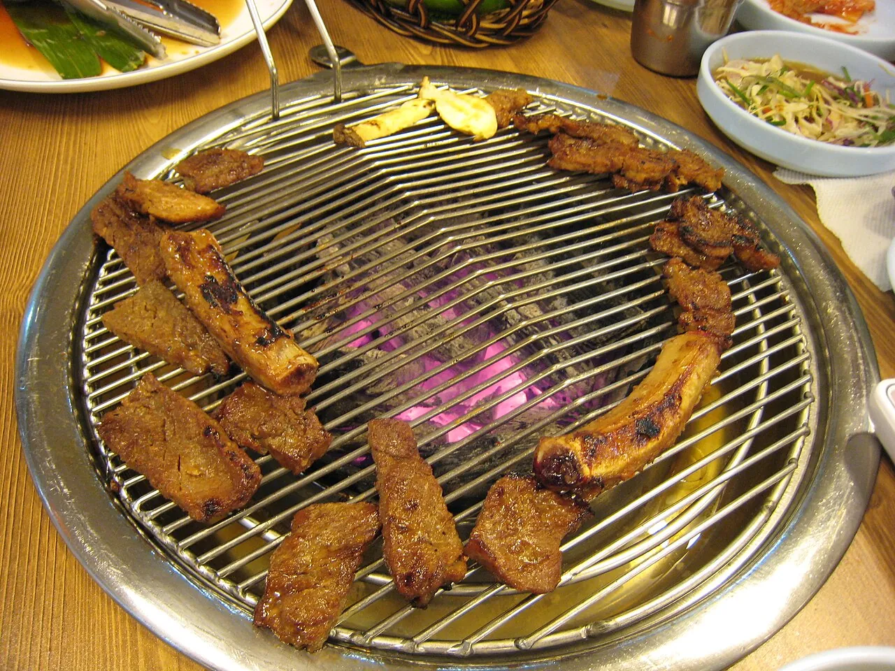 Marinated pork ribs grilling on a round charcoal table grill with side dishes around it
