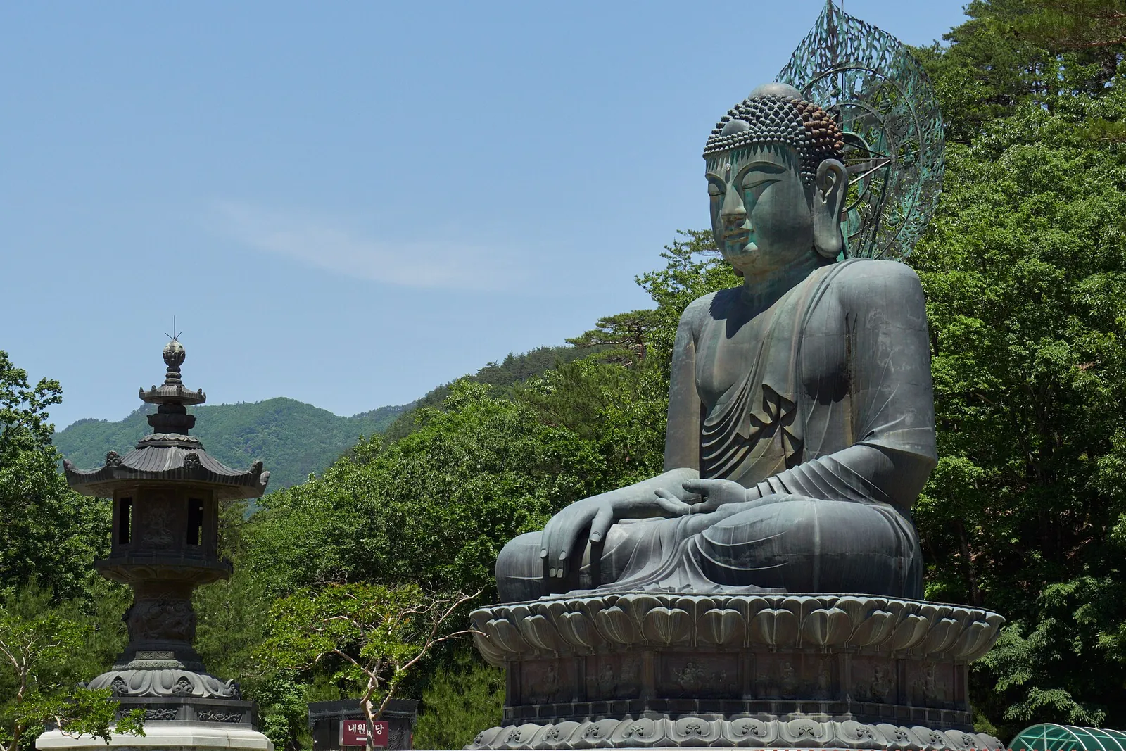 A large bronze seated Buddha statue beside a stone lantern at Sinheungsa temple, with forested hills behind