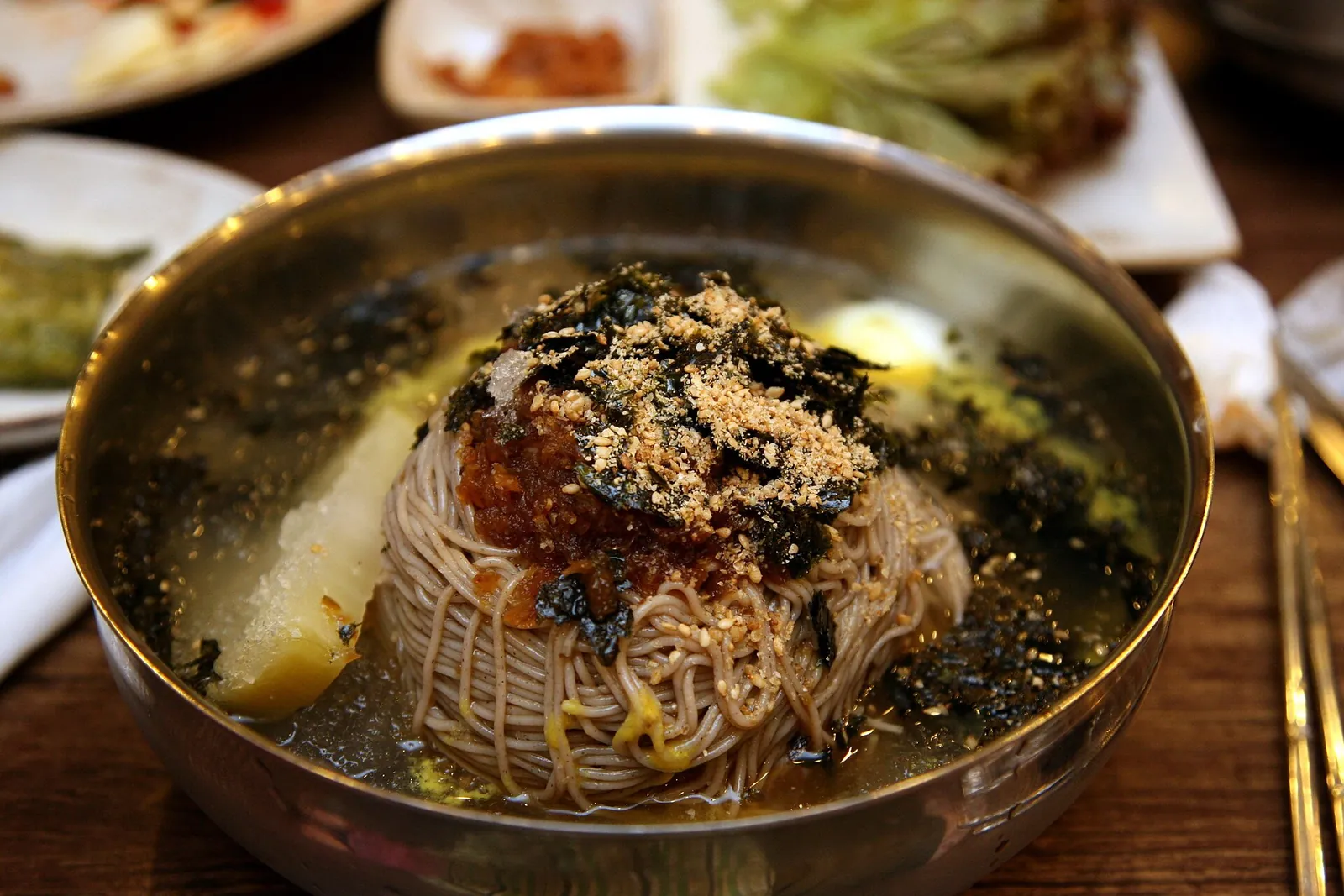 A metal bowl of makguksu, buckwheat noodles in cold broth topped with chilli sauce, seaweed and sesame