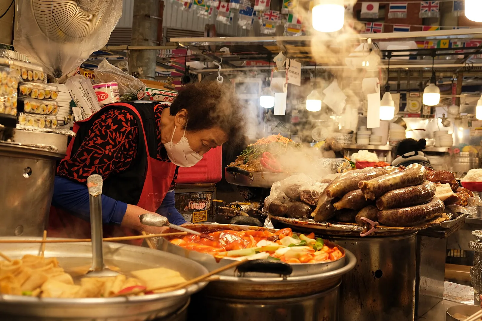 A vendor in a mask serving from steaming trays of fried pancakes and other dishes at a Gwangjang Market stall